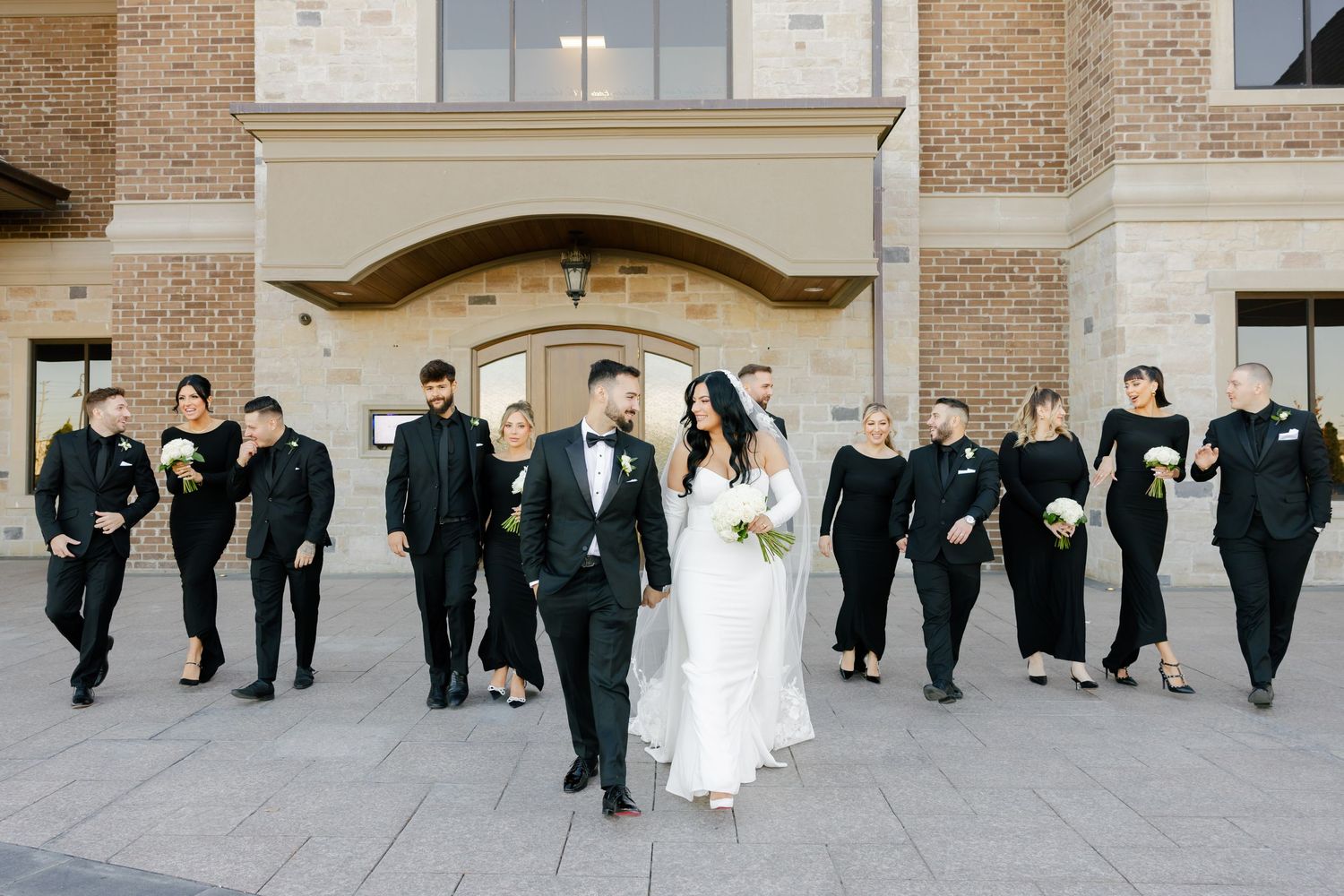 Wedding party in black and white attire walks together outside a brick building.