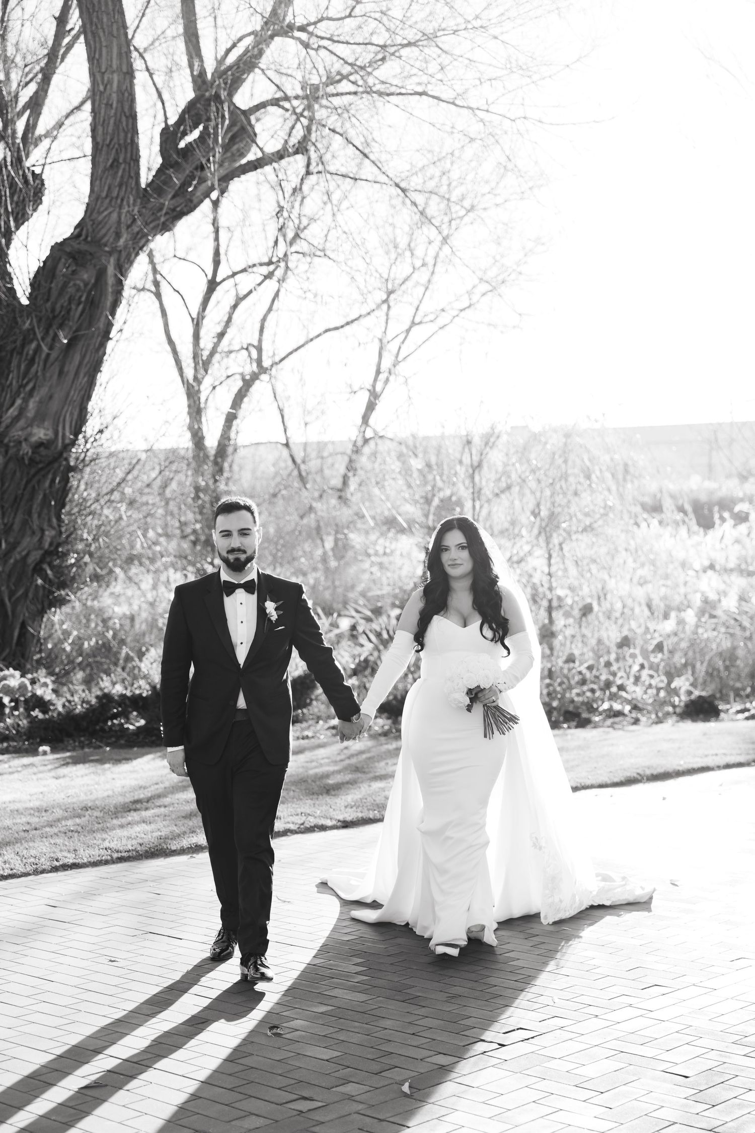 Black and white wedding photo of couple walking along brick pathway under bare trees.