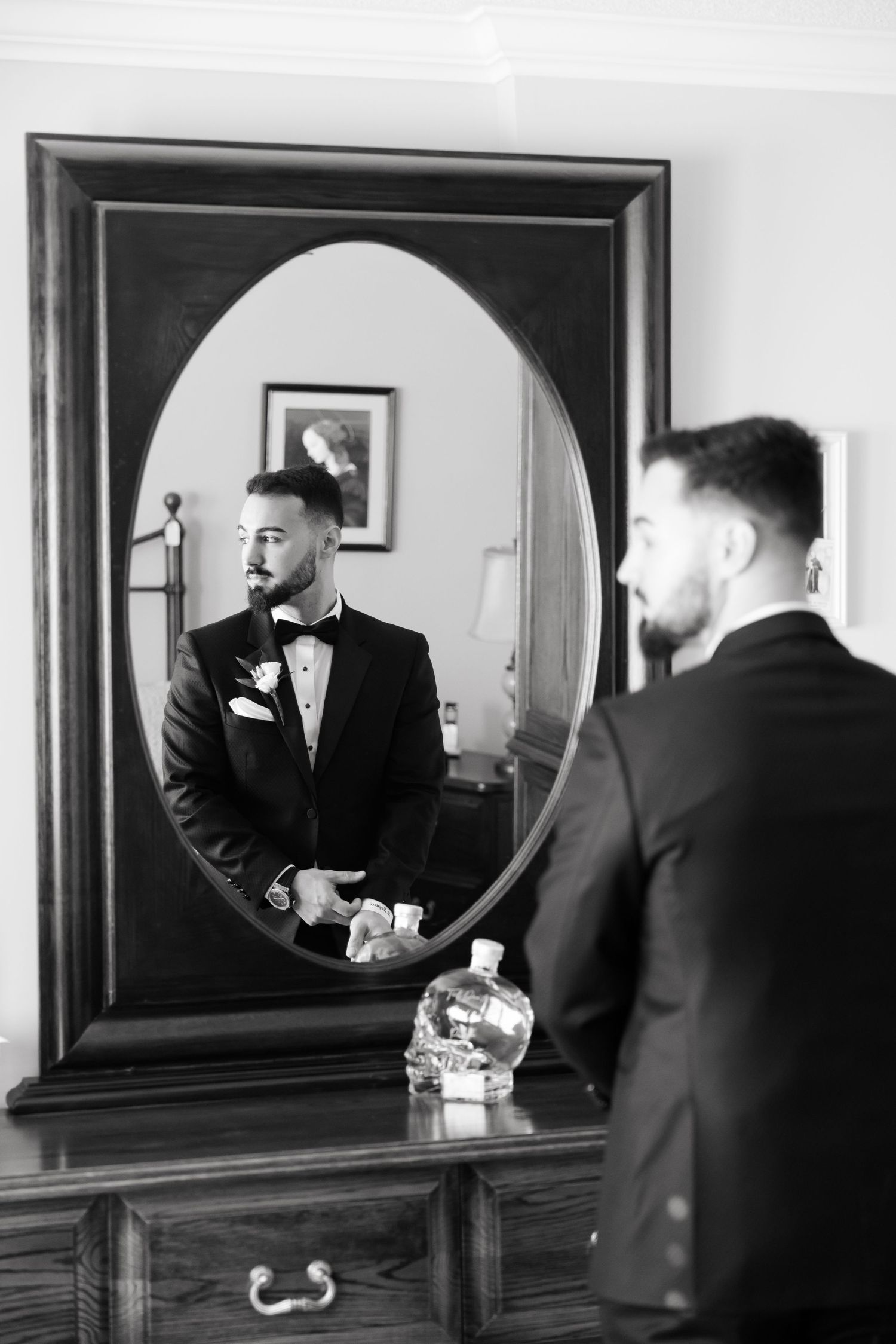 A black and white portrait of a groom checking his appearance in an oval mirror.