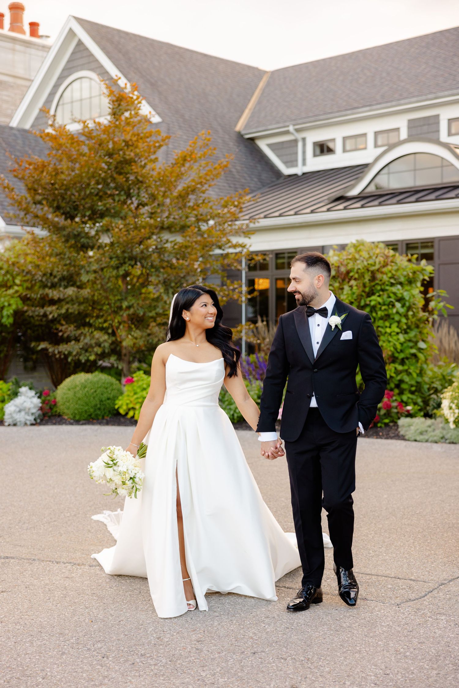 A wedding couple walks hand in hand on a path outside a gray house with autumn trees in the background.