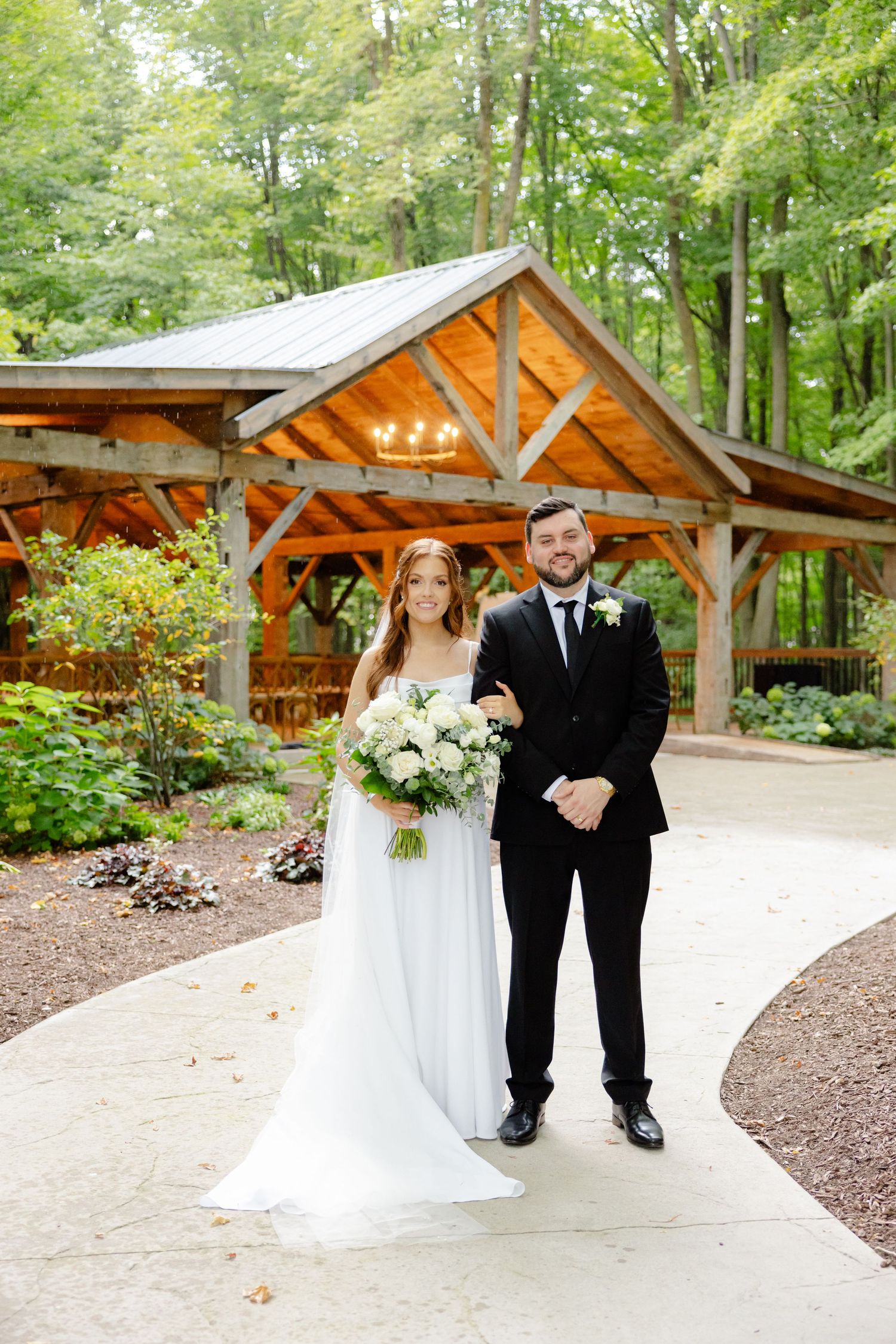Wedding portrait in front of rustic wooden pavilion surrounded by forest greenery.