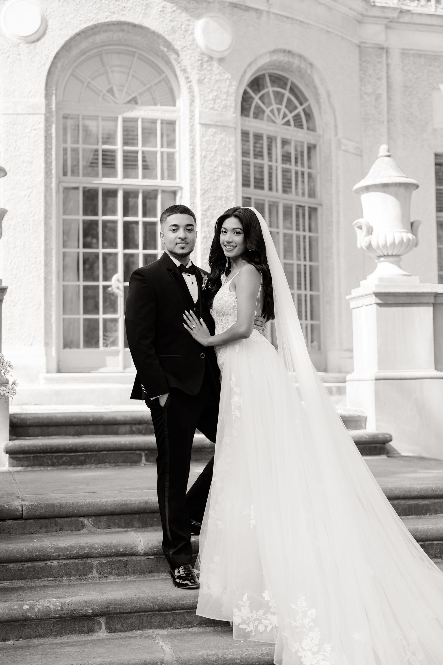A black and white wedding portrait captured on the steps of a historic mansion with arched windows.