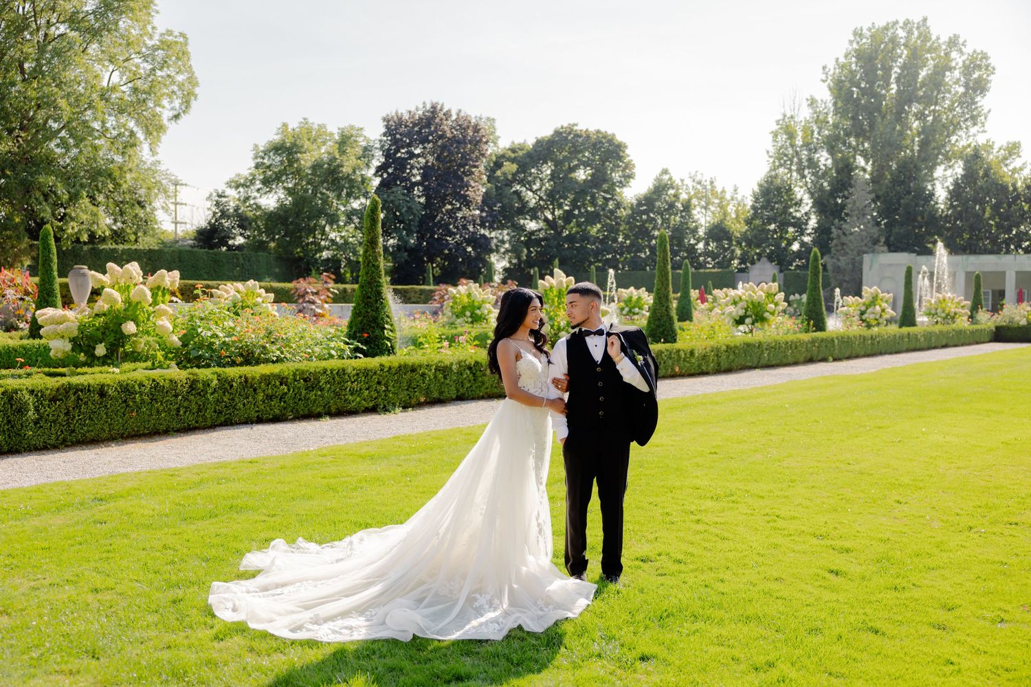 A wedding couple poses together on a lush green lawn with formal gardens and topiaries in the background.