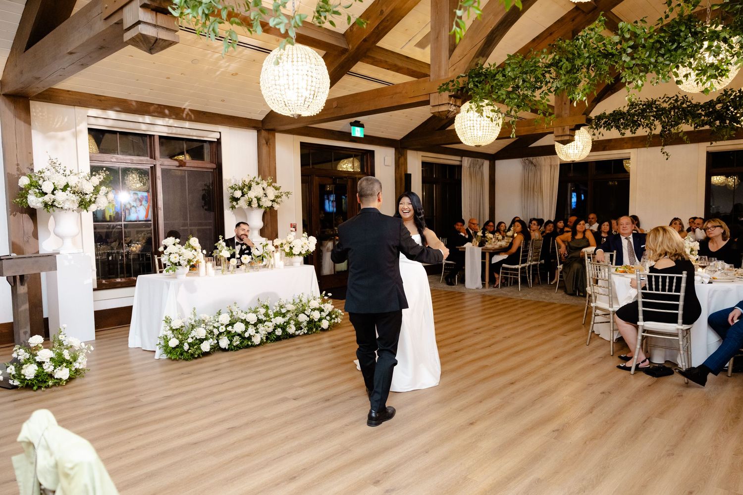 Reception hall with guests watching couple dance under crystal chandeliers and floral arrangements.