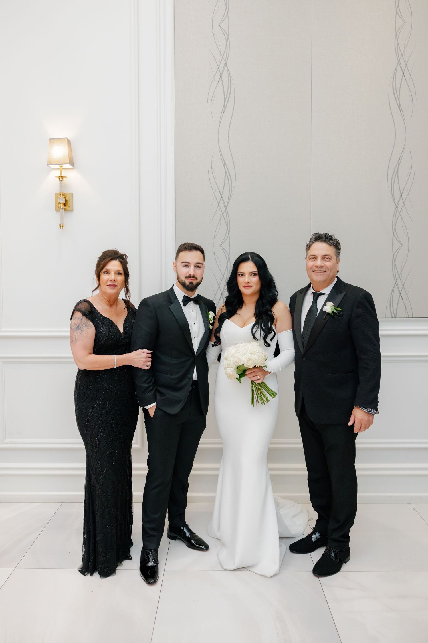 Formal wedding party portrait in black attire against a white wall with elegant sconce lighting.