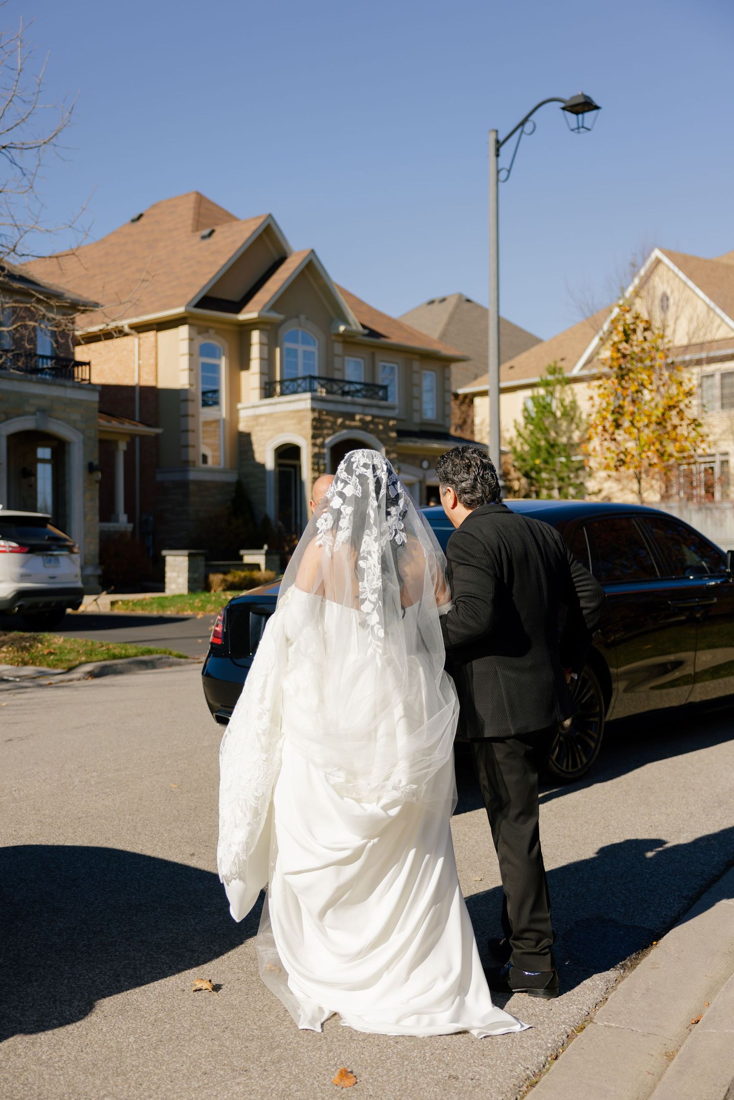 Bride in flowing white dress and veil walking toward luxury car on sunny suburban street.