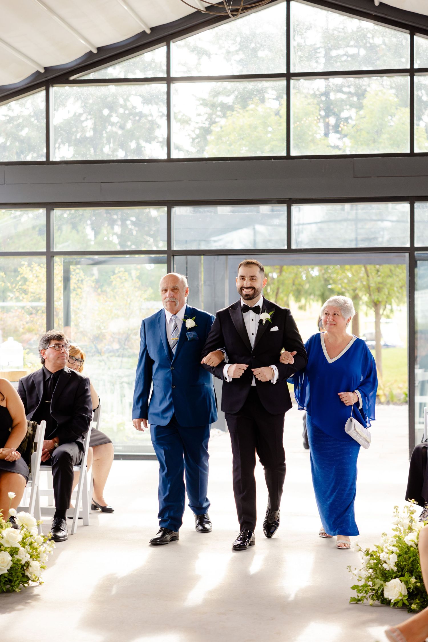 Wedding party members walk down the aisle in a modern venue with large windows and floral decorations.