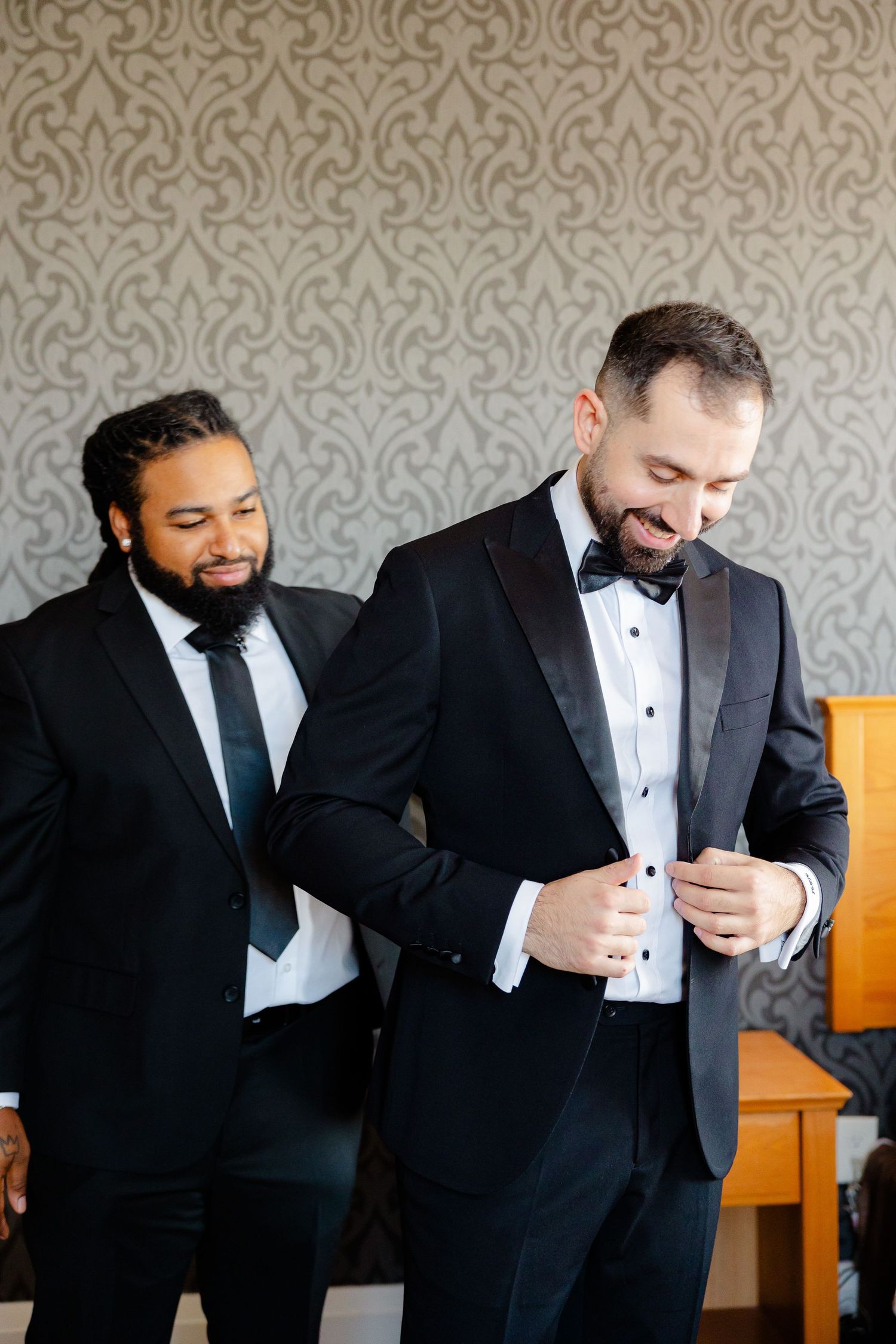 Two people in formal attire sharing a lighthearted moment against damask wallpaper.