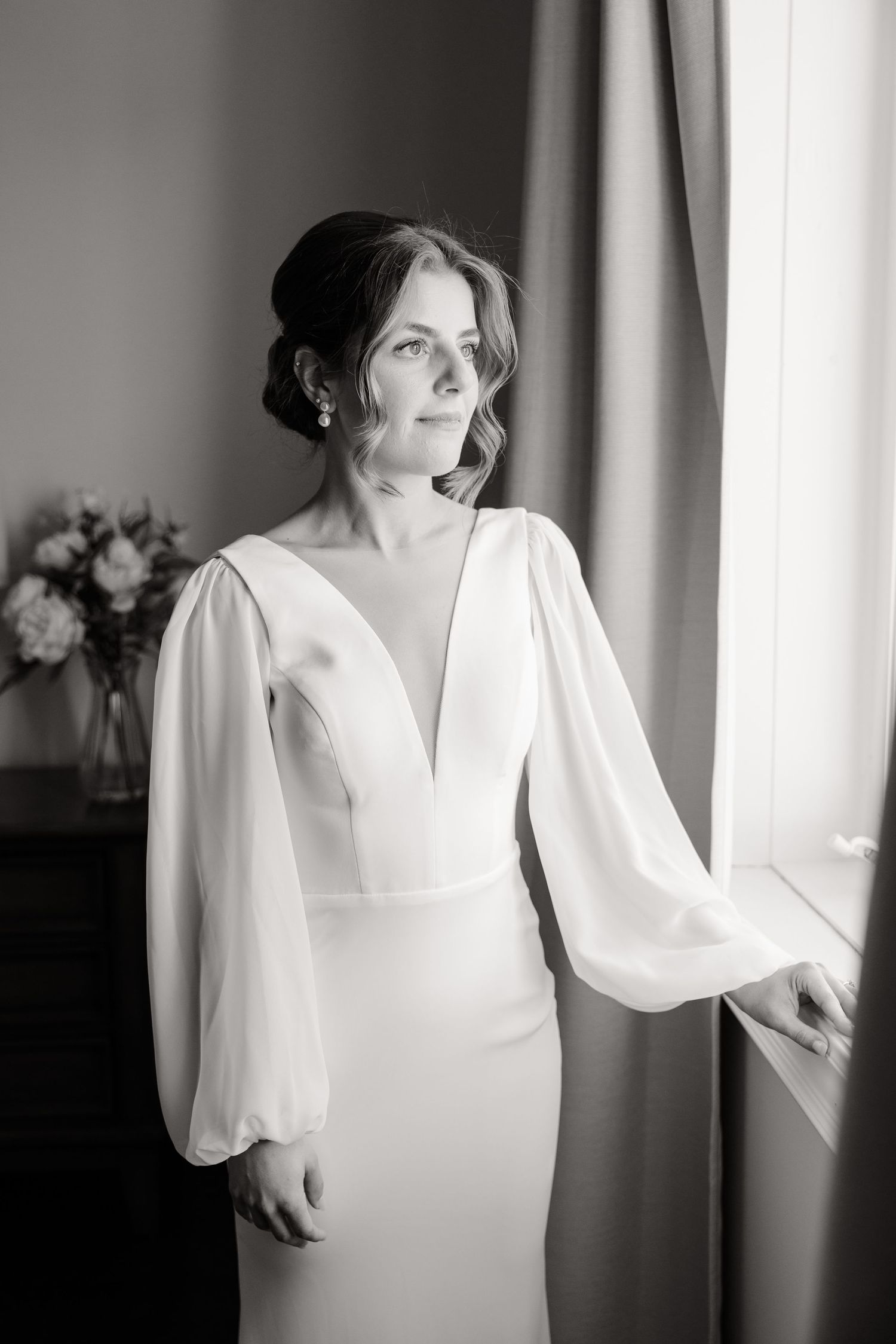 Black and white portrait of an elegant bridal gown with dramatic bishop sleeves beside a window.