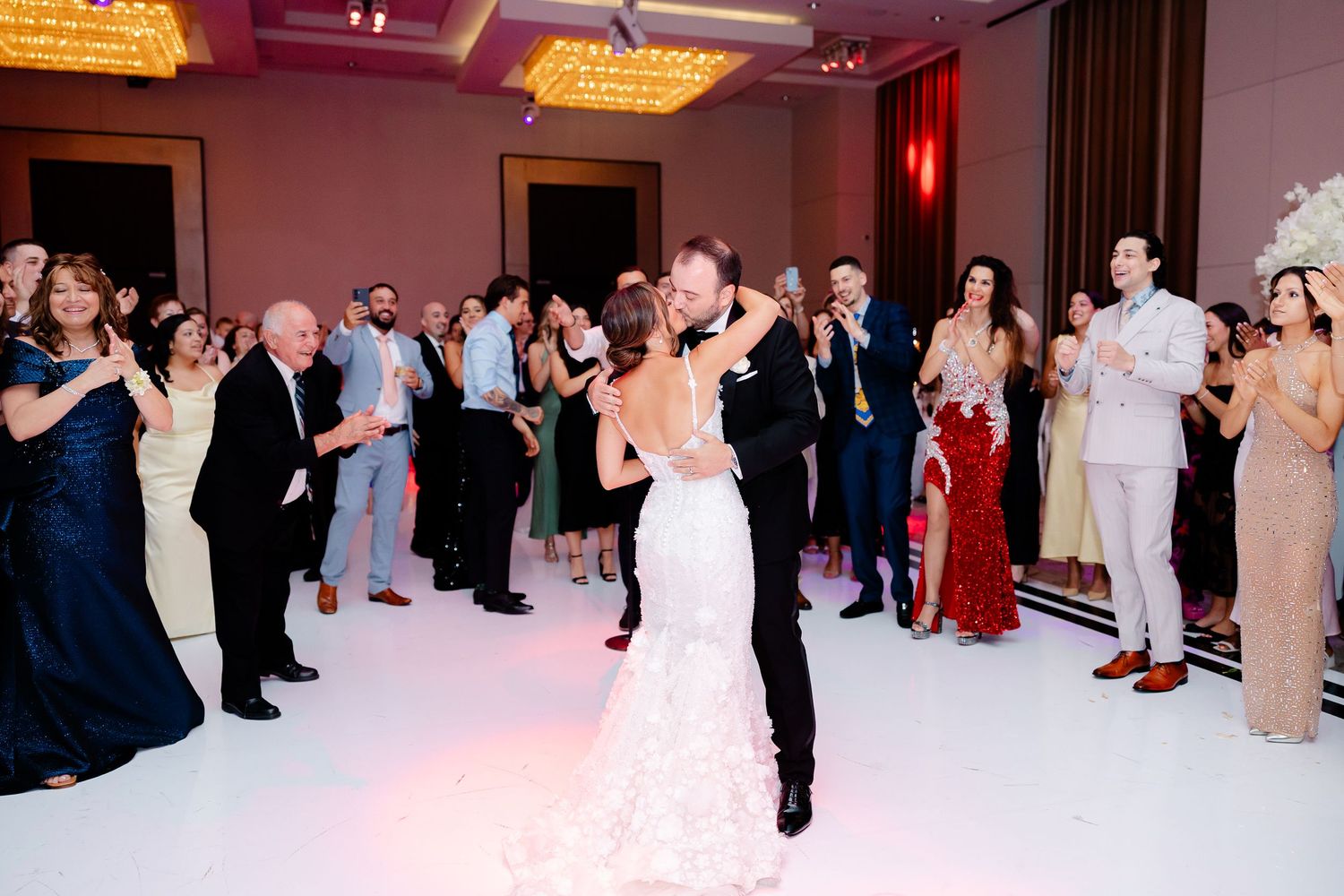 Guests gather around a dance floor at an elegant wedding reception with pink lighting and chandeliers overhead.