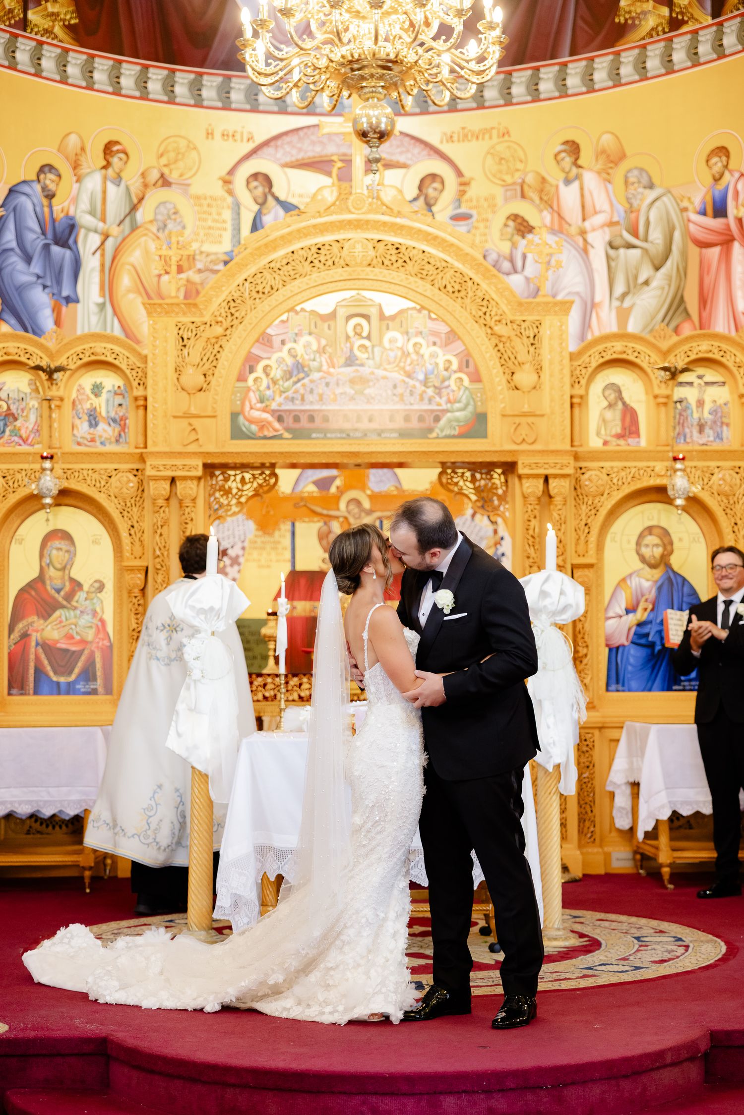 Wedding ceremony taking place in an ornate orthodox church with golden murals and religious icons decorating the walls.