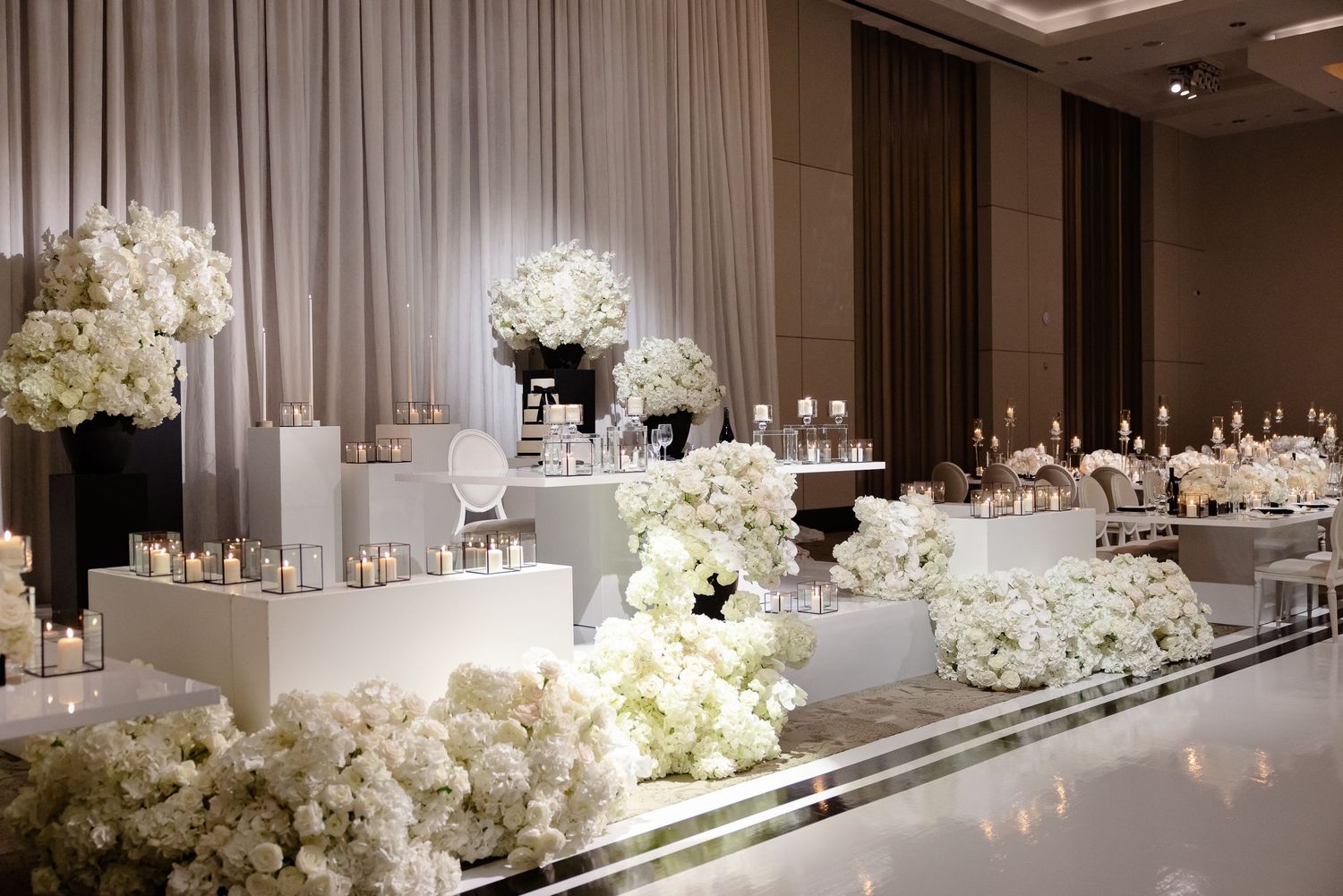 Elegant wedding reception setup with white floral arrangements and black vases on mirrored tables in a modern ballroom.