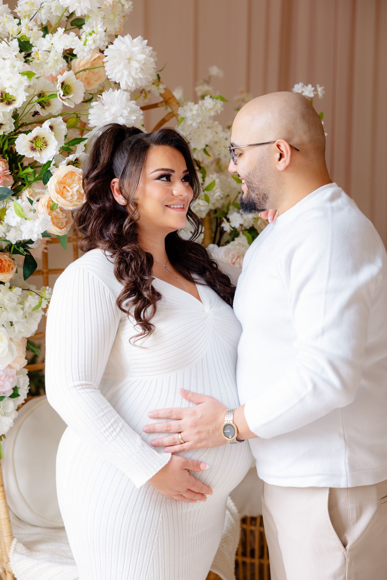 An expecting couple in matching white outfits embraces in front of a floral backdrop.