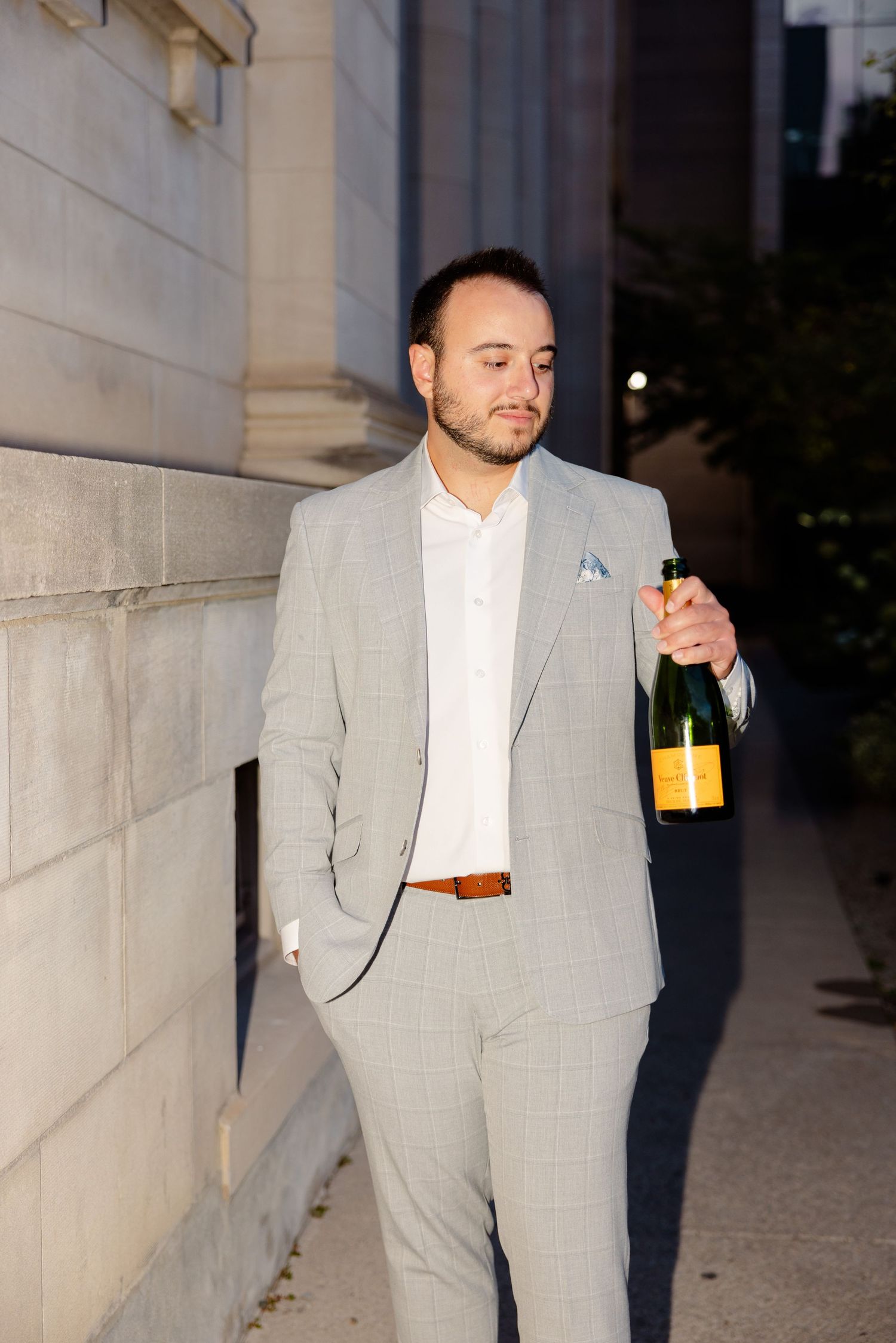 Well-dressed man in light gray suit holds champagne bottle while standing against stone pillar.