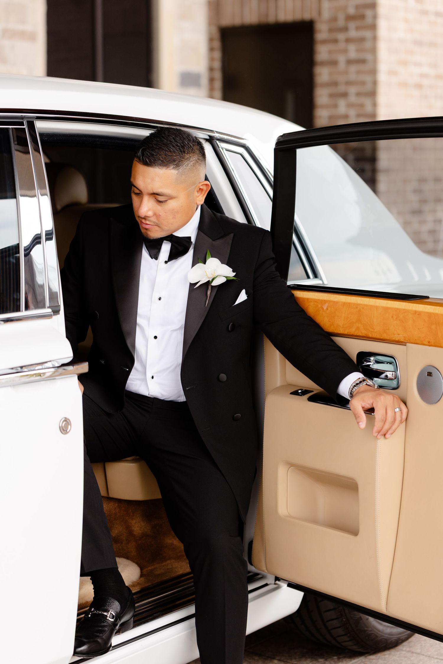 Groom in black tuxedo entering classic vintage white car with tan leather interior for wedding transportation.