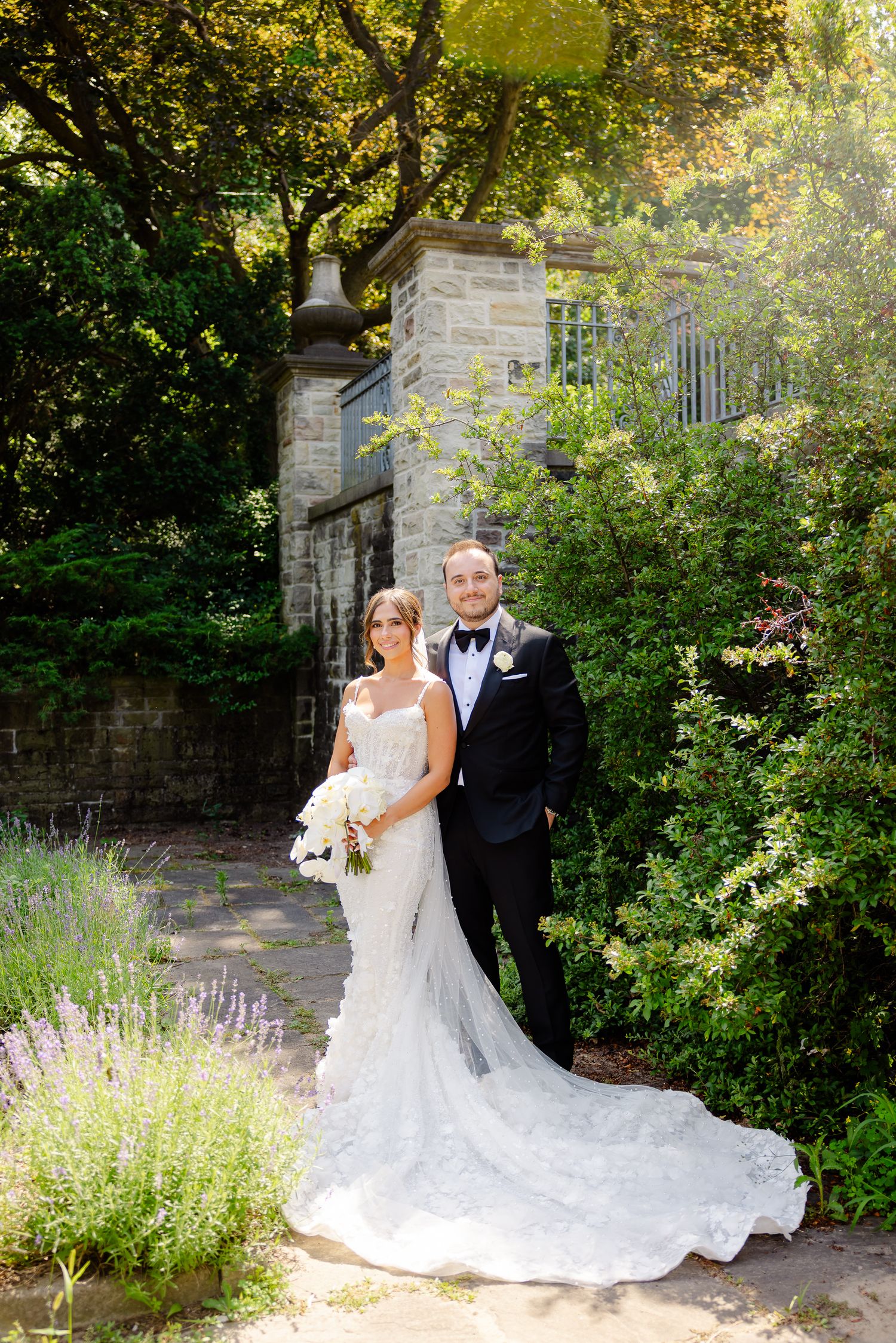 A bride in a white lace gown with long train and groom in black tuxedo pose in a sunlit garden with stone pillars.
