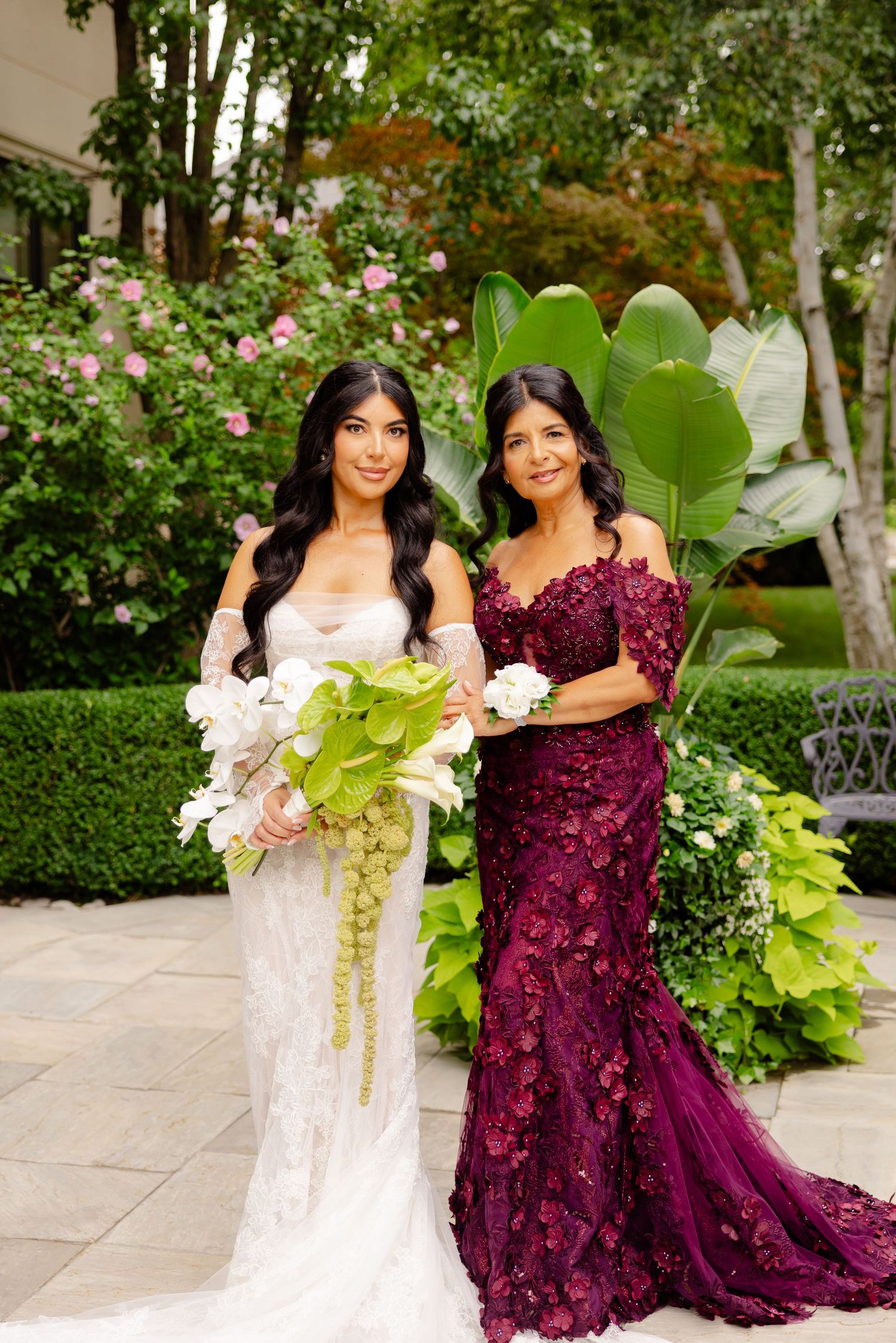 Wedding party members in white and burgundy dresses pose together on an elegant stone patio with garden backdrop.