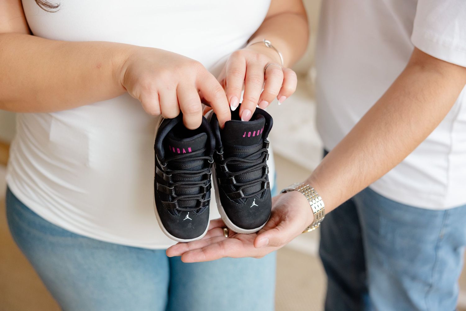 Hands hold a tiny pair of black baby sneakers against a white background.