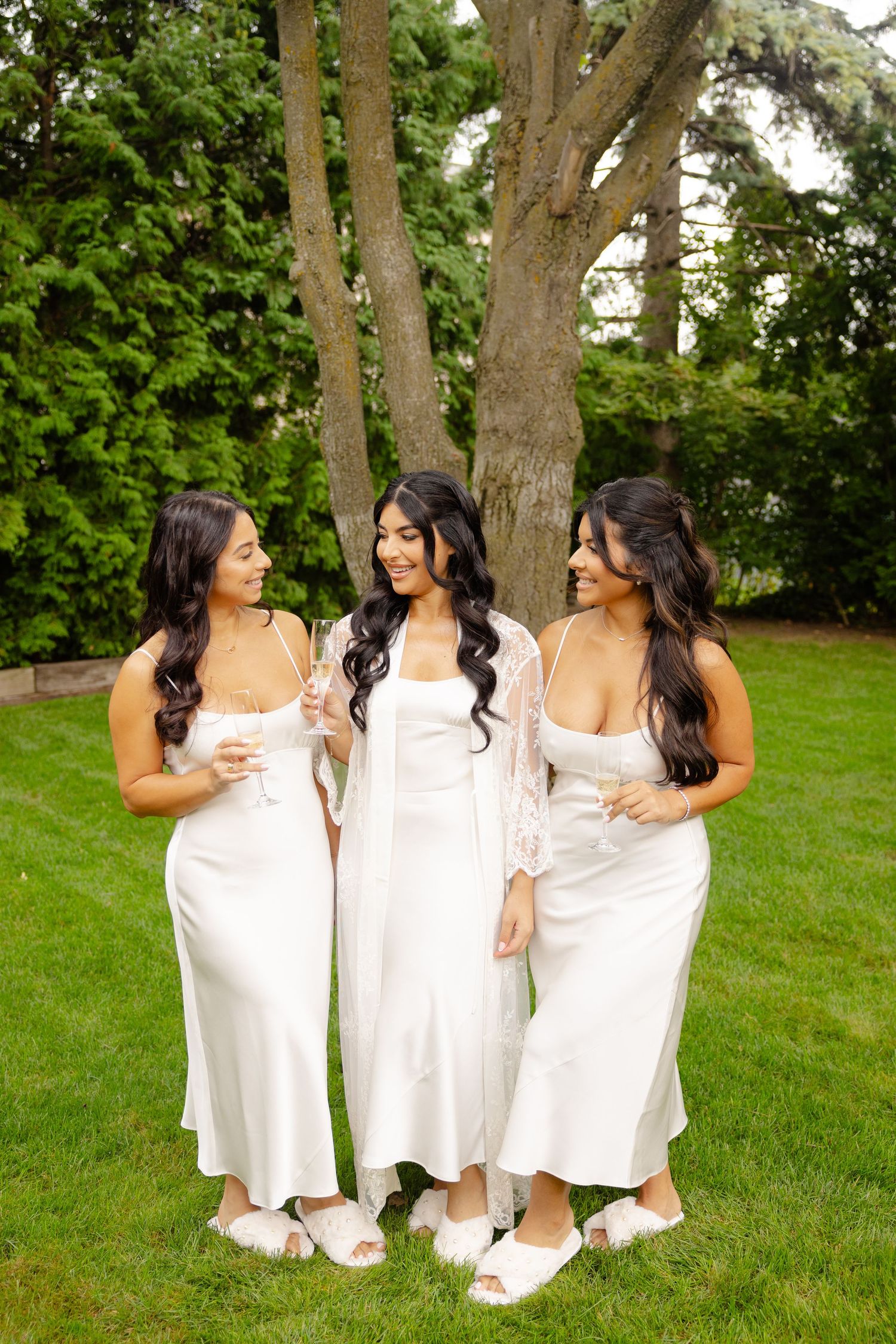 Three women in flowing white dresses sit close together on green grass with trees in the background.