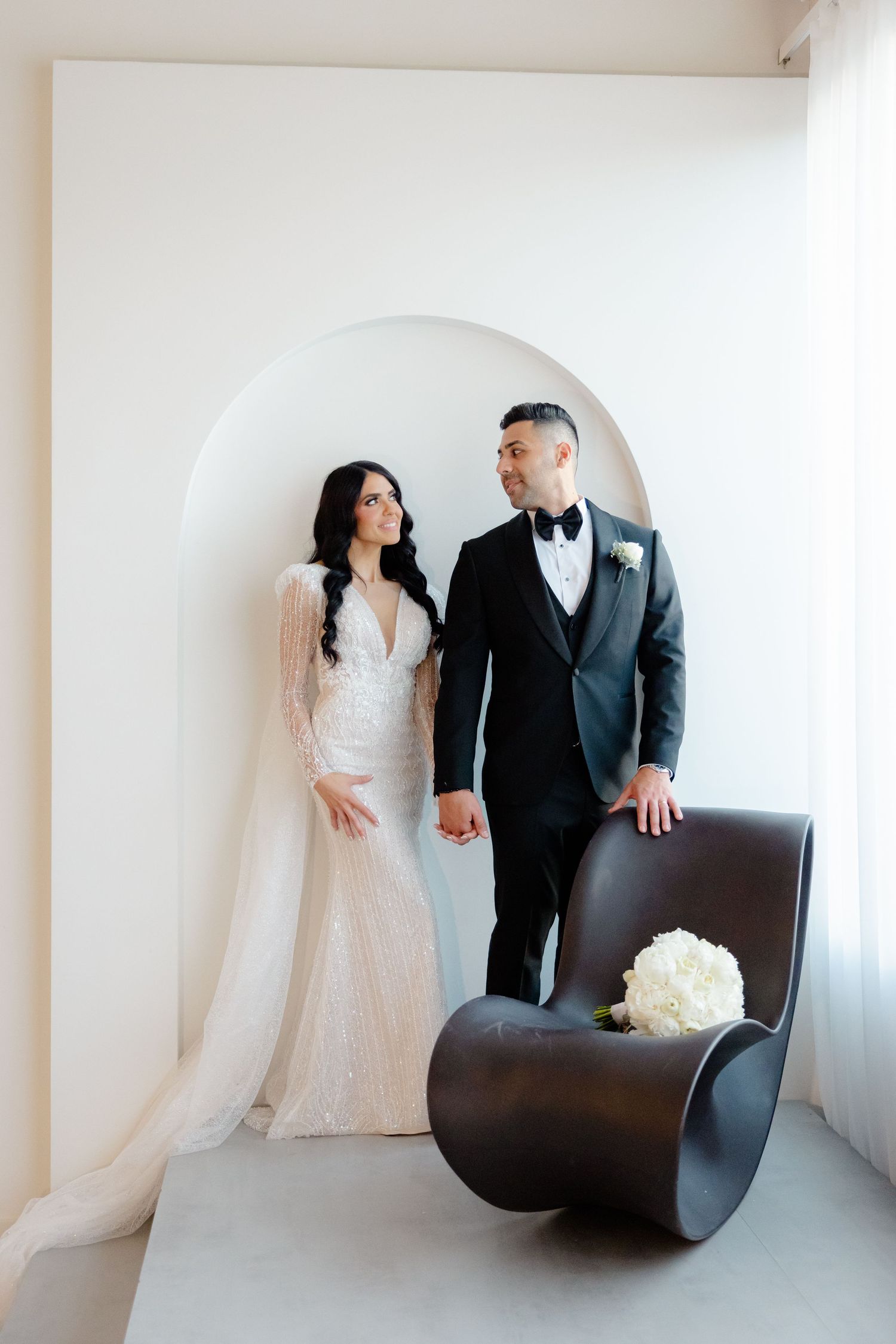 Stylish bride and groom standing in minimalist white setting with modern curved chair.