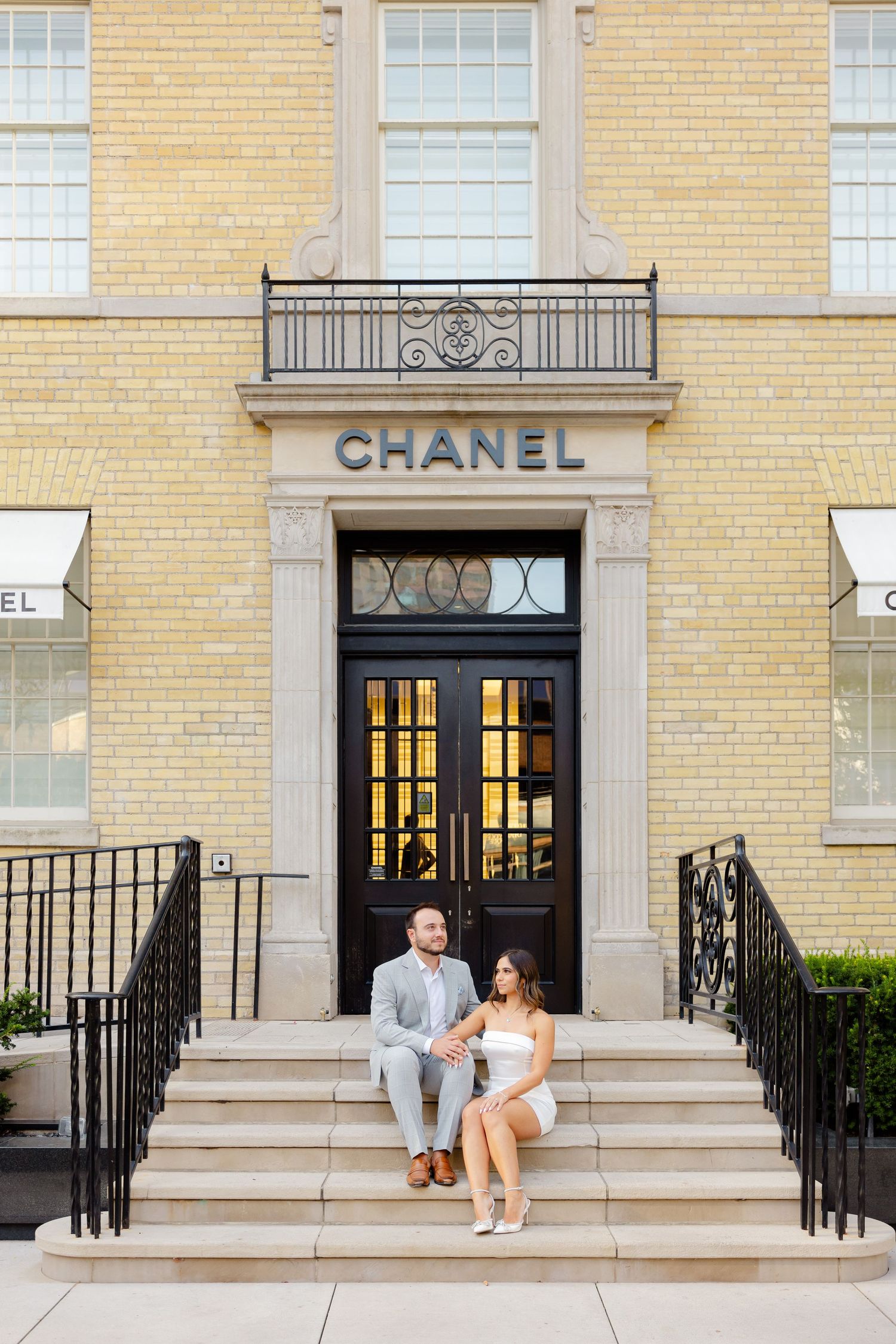 Couple relaxing on stone steps of luxury Chanel boutique with ornate doorway and yellow brick facade.
