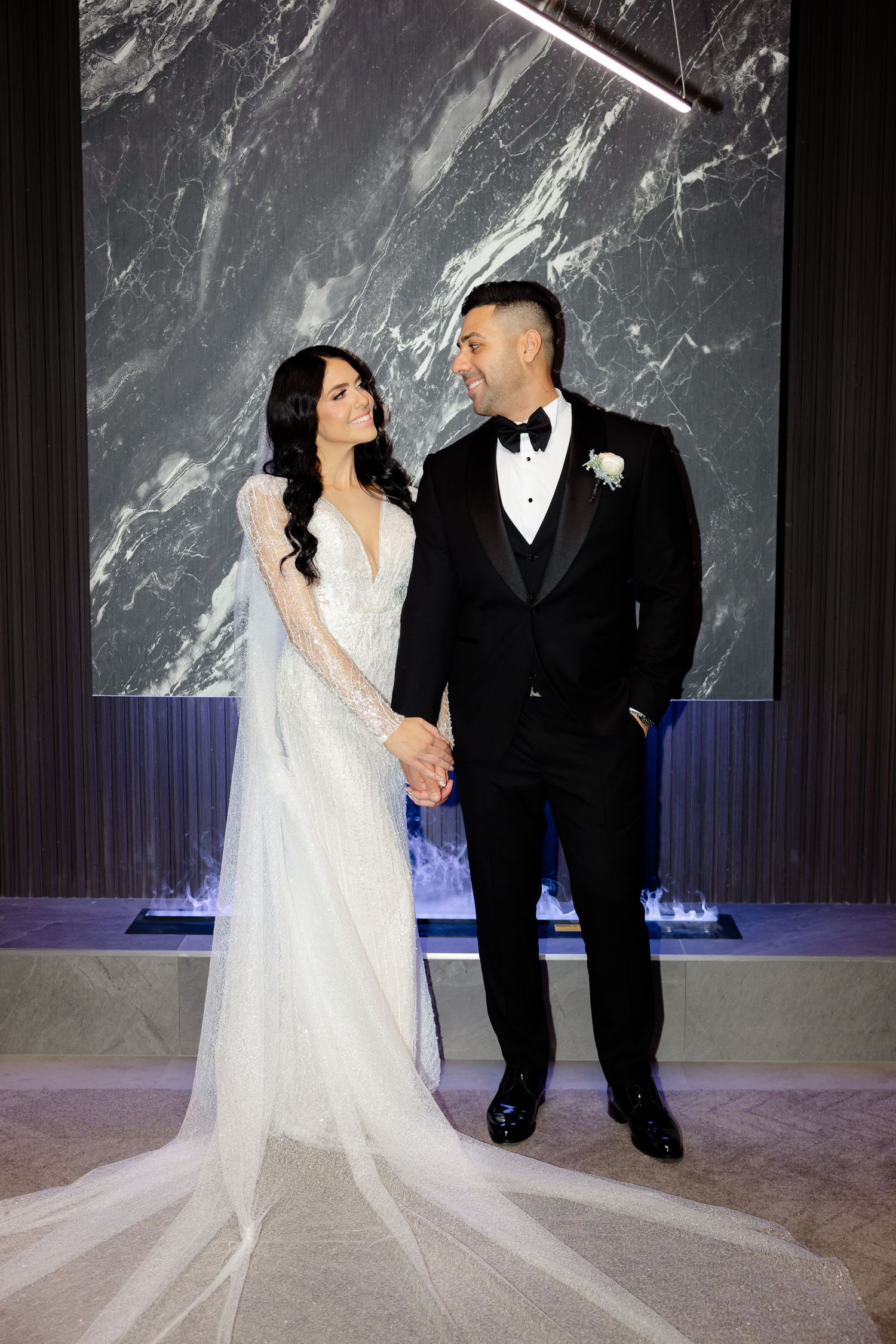 Newlyweds share intimate moment against marble wall with geometric lighting fixture.