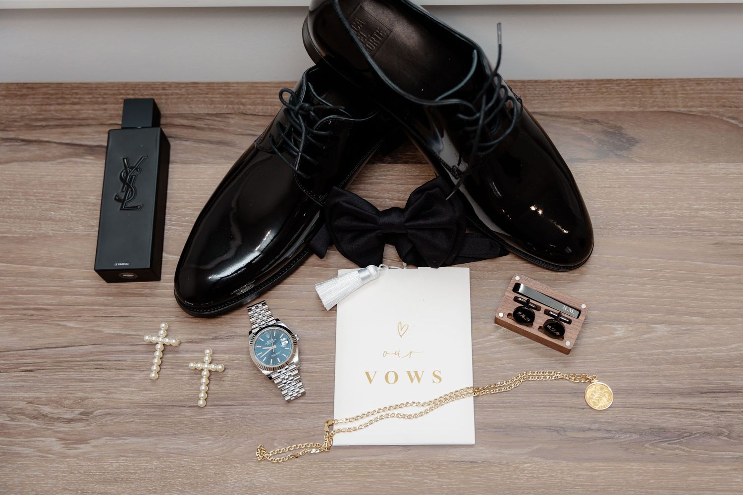 Groom's wedding accessories including shoes, crosses, watch and vows card arranged on wooden floor.