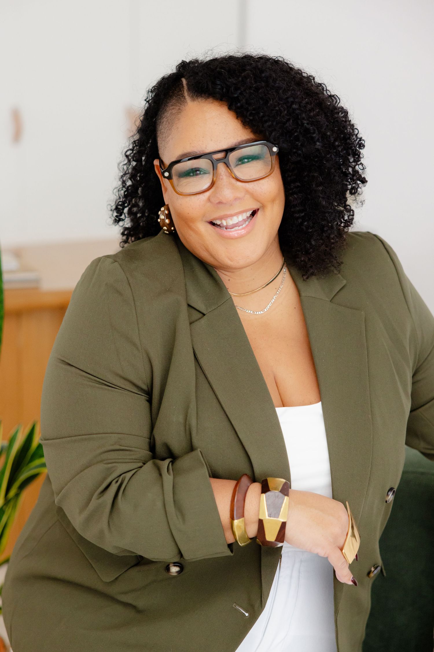 Professional portrait of someone in a stylish olive blazer and glasses smiling warmly against a light background.
