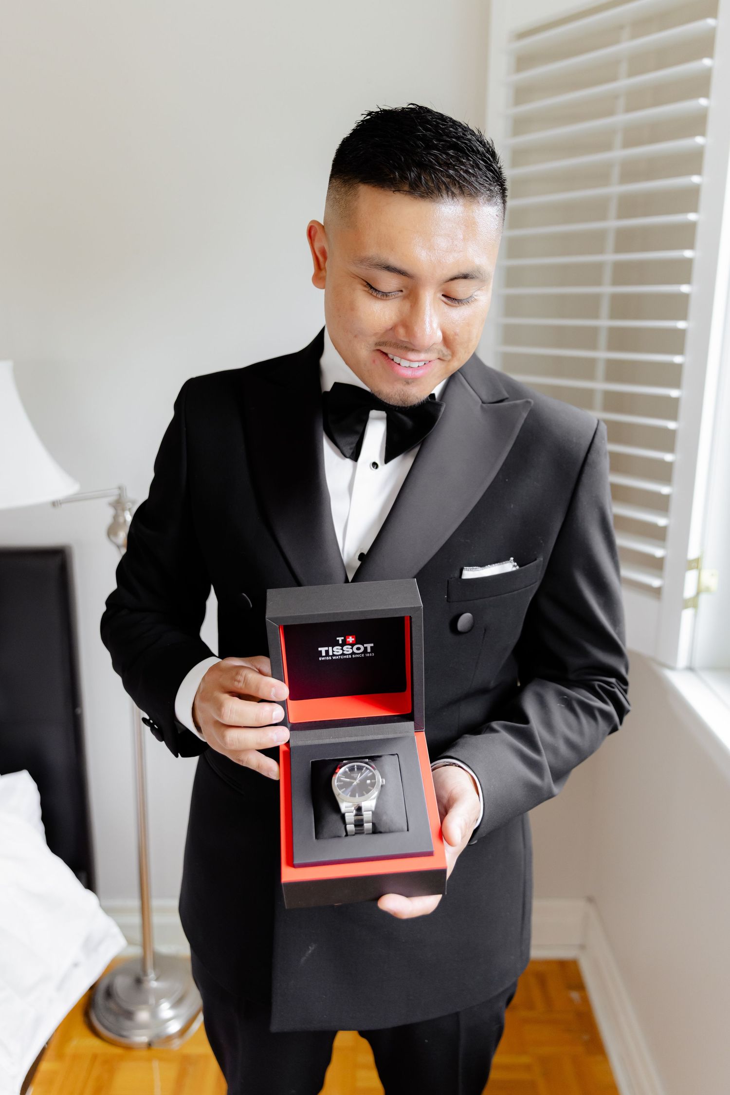 A groom wearing a classic black tuxedo smiles while opening a gift box containing a watch in a well-lit room.