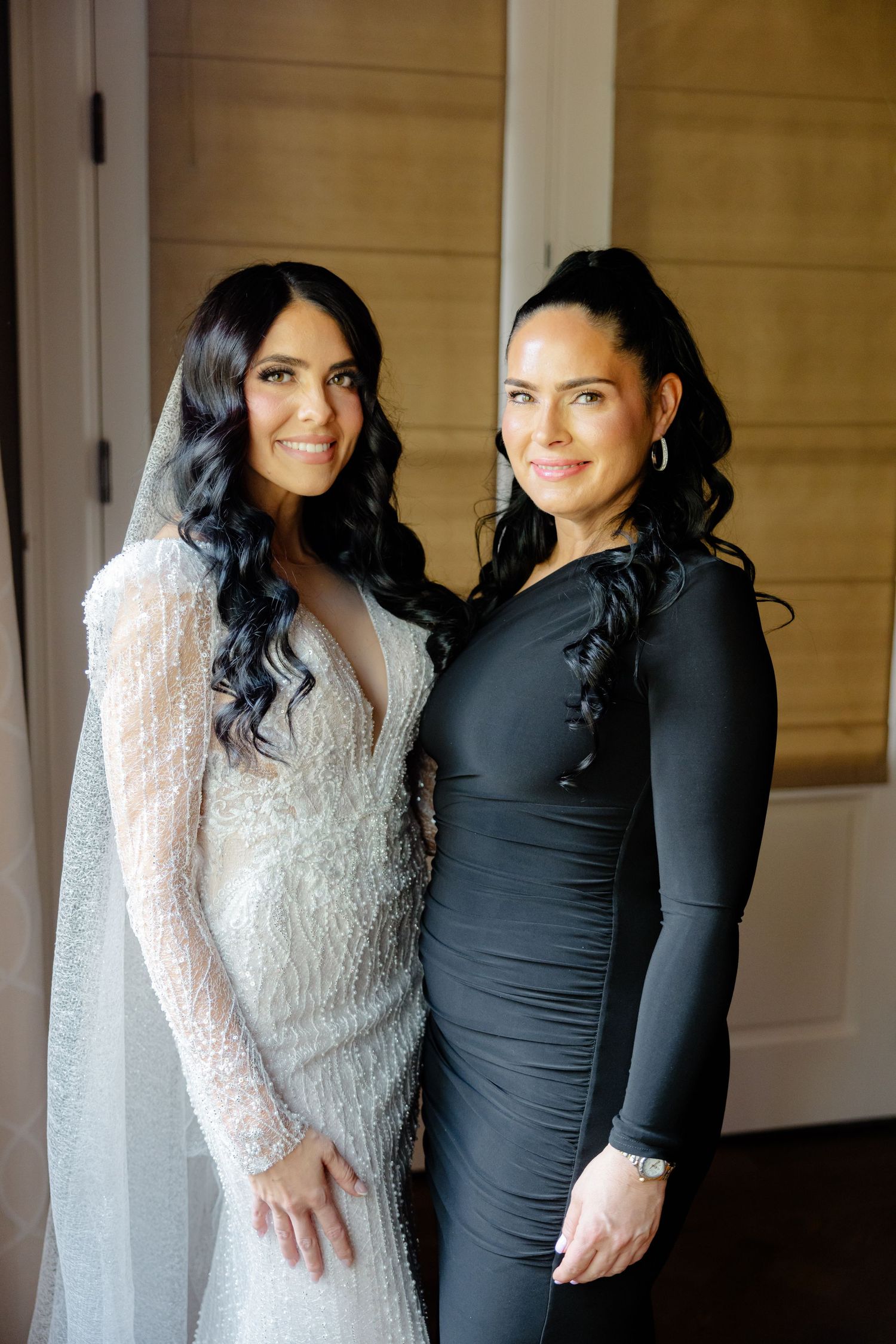 A bride in a white lace gown stands with a woman in a fitted black dress against a wooden wall.
