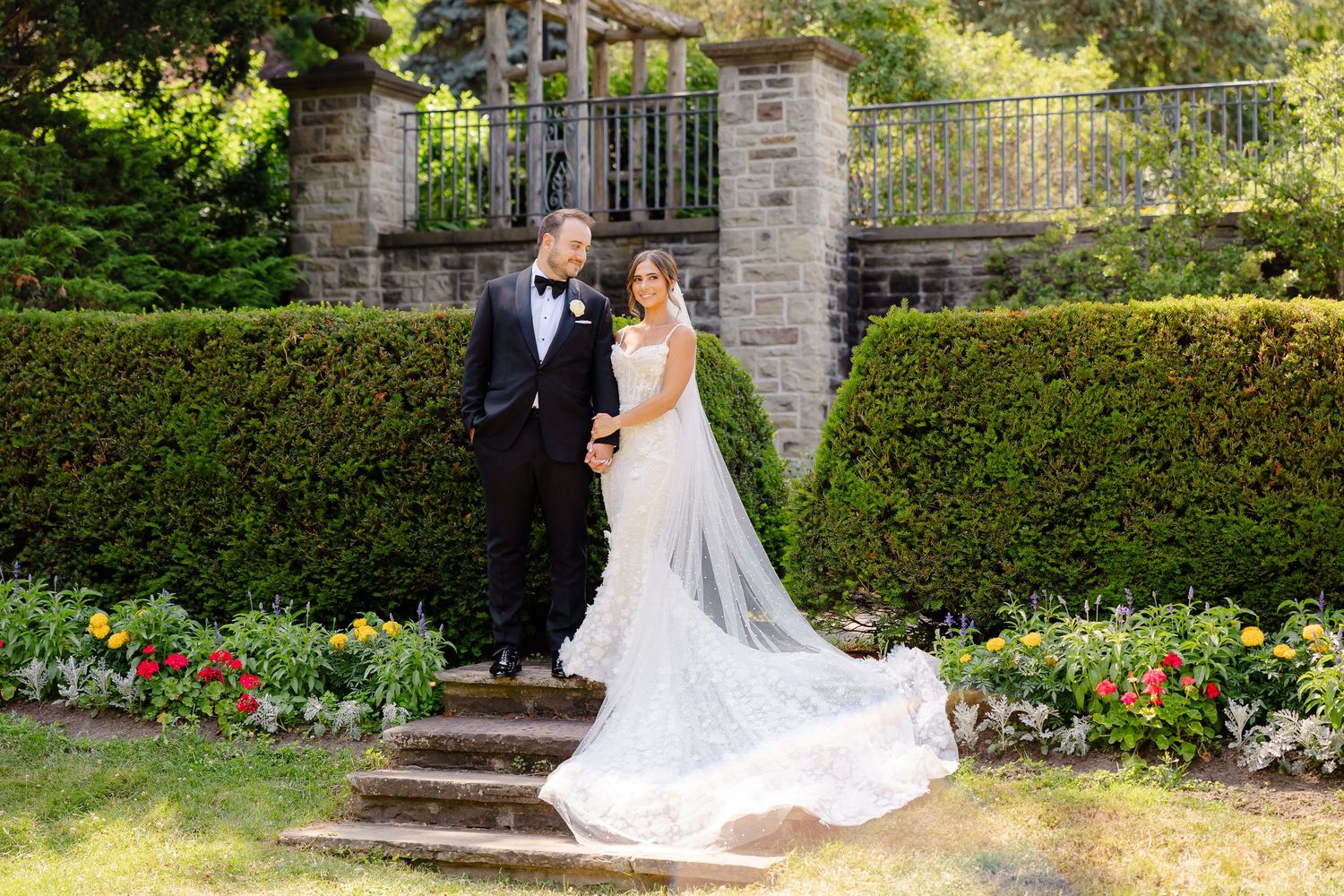 A bride and groom stand on garden steps surrounded by manicured hedges and colorful flowers at a formal estate.
