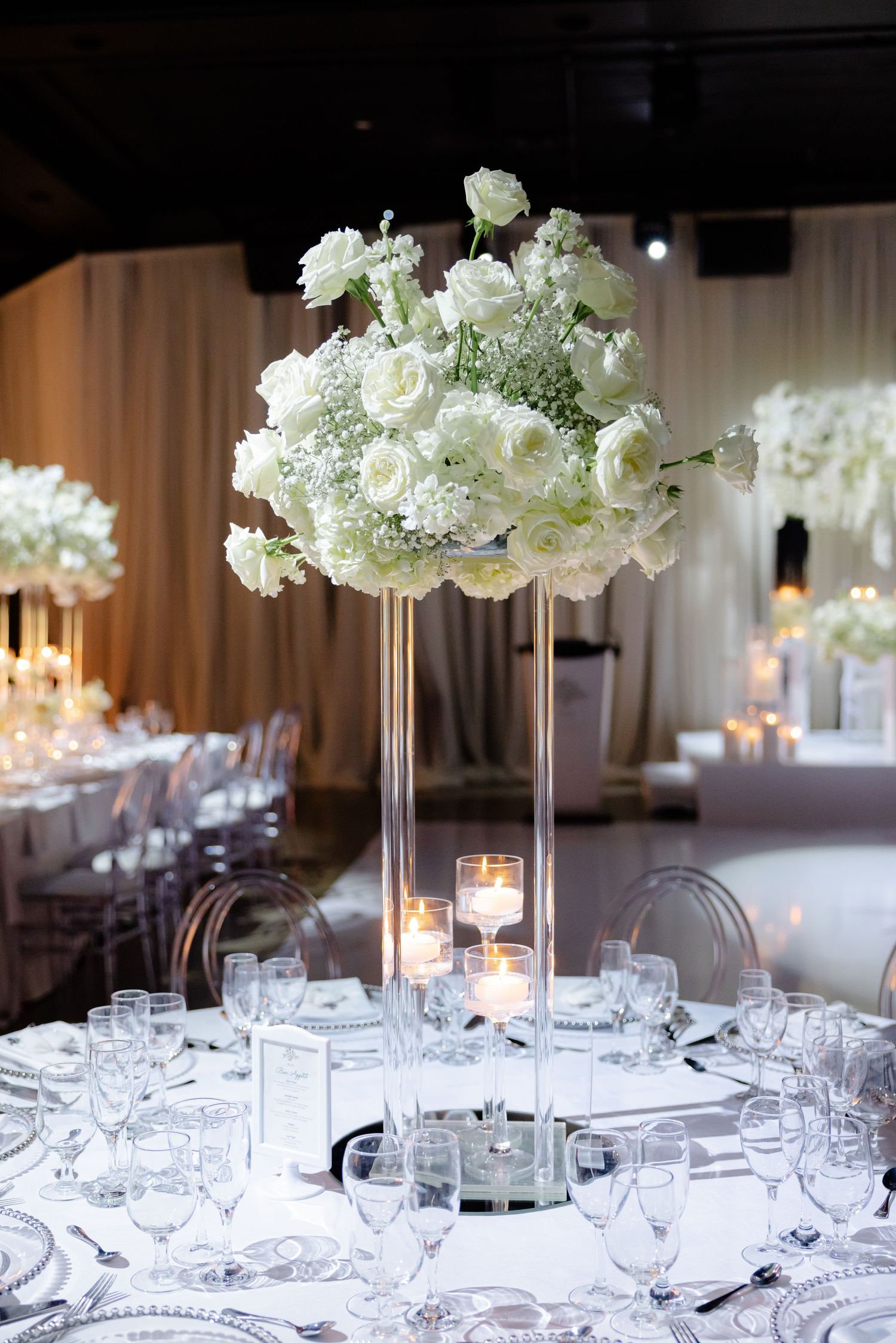 White floral centerpiece on tall glass pedestal adorning round reception table with crystal place settings.