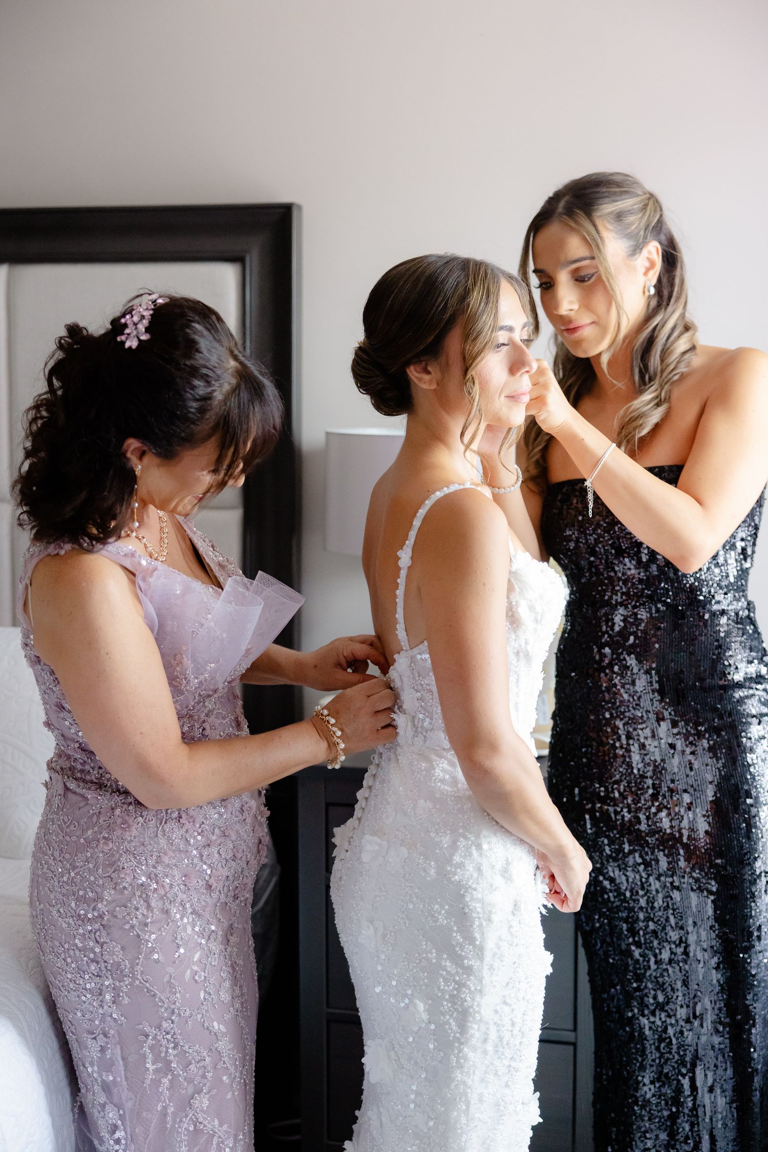 Bridesmaids help bride get ready, one wearing lavender and another in black sequin gown adjust white wedding dress.