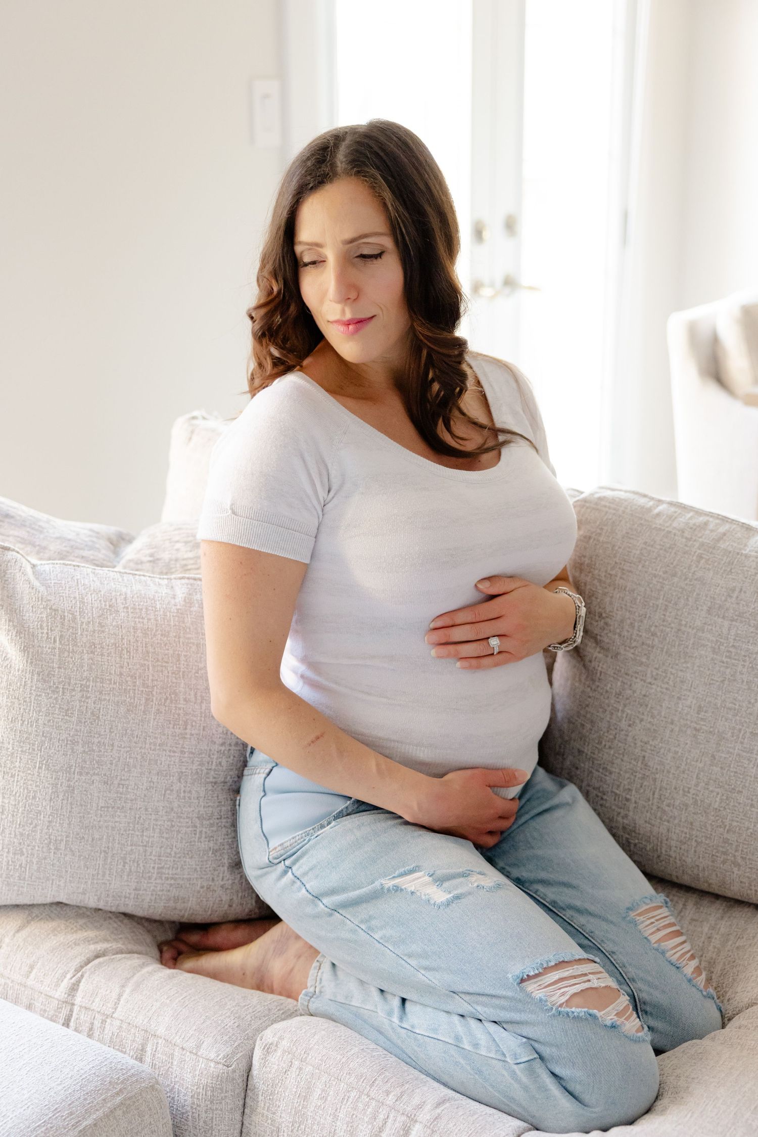 A pregnant woman in a white shirt and jeans sits on a couch, gently cradling her belly with her eyes closed.