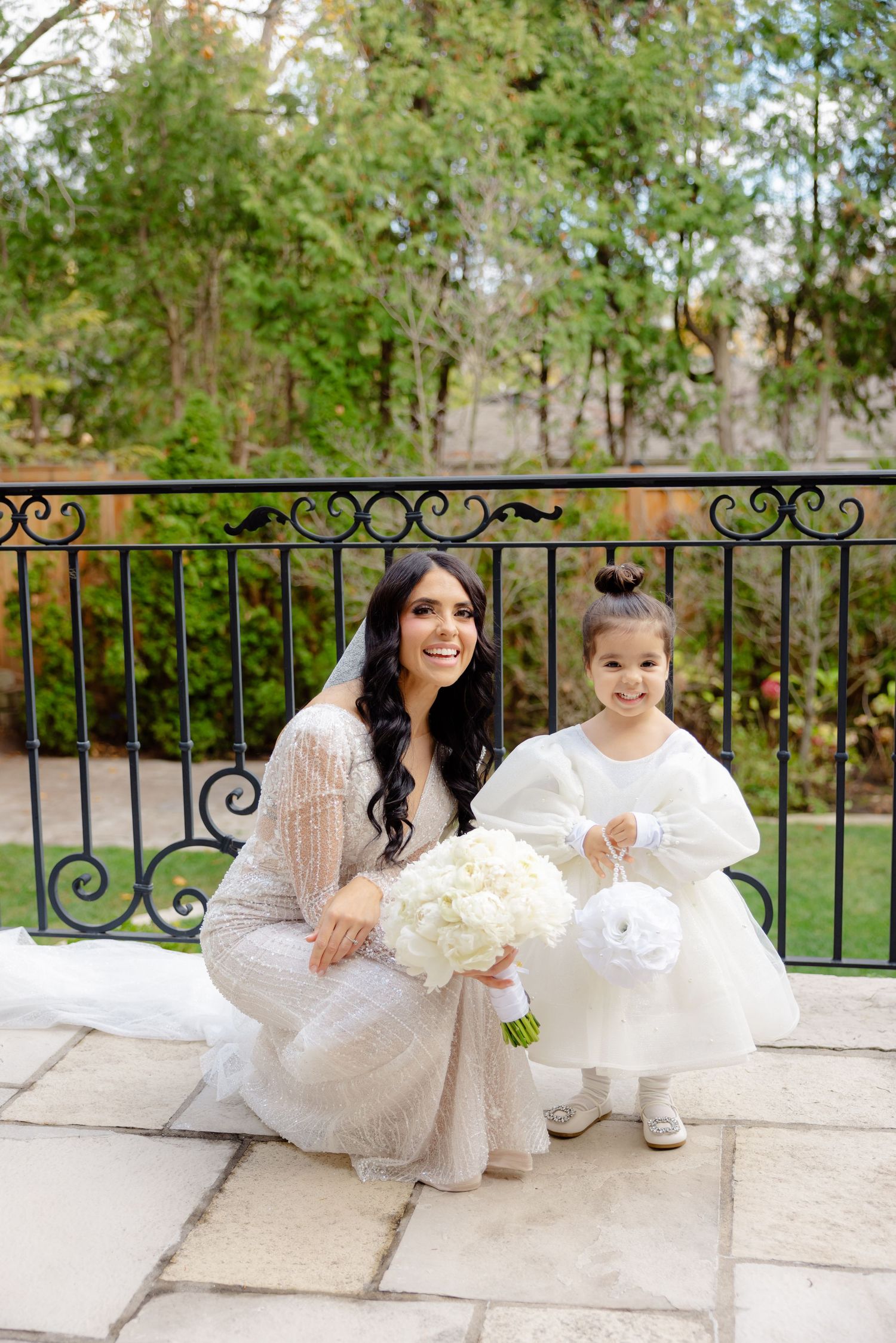 Bride and flower girl share bright smiles while posing together on stone terrace.