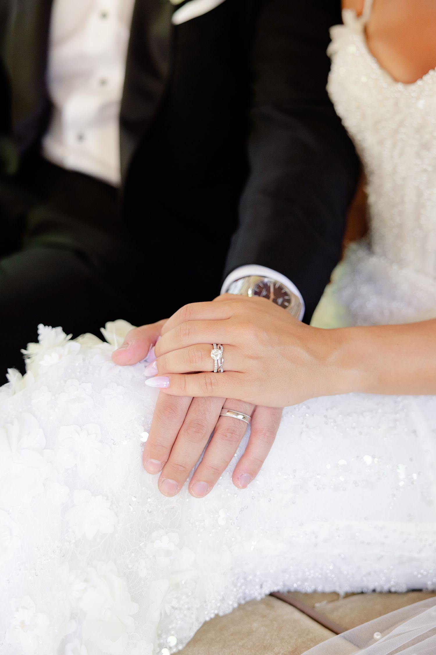 A close-up of hands showing wedding rings against white lace dress fabric and formal black suit attire.