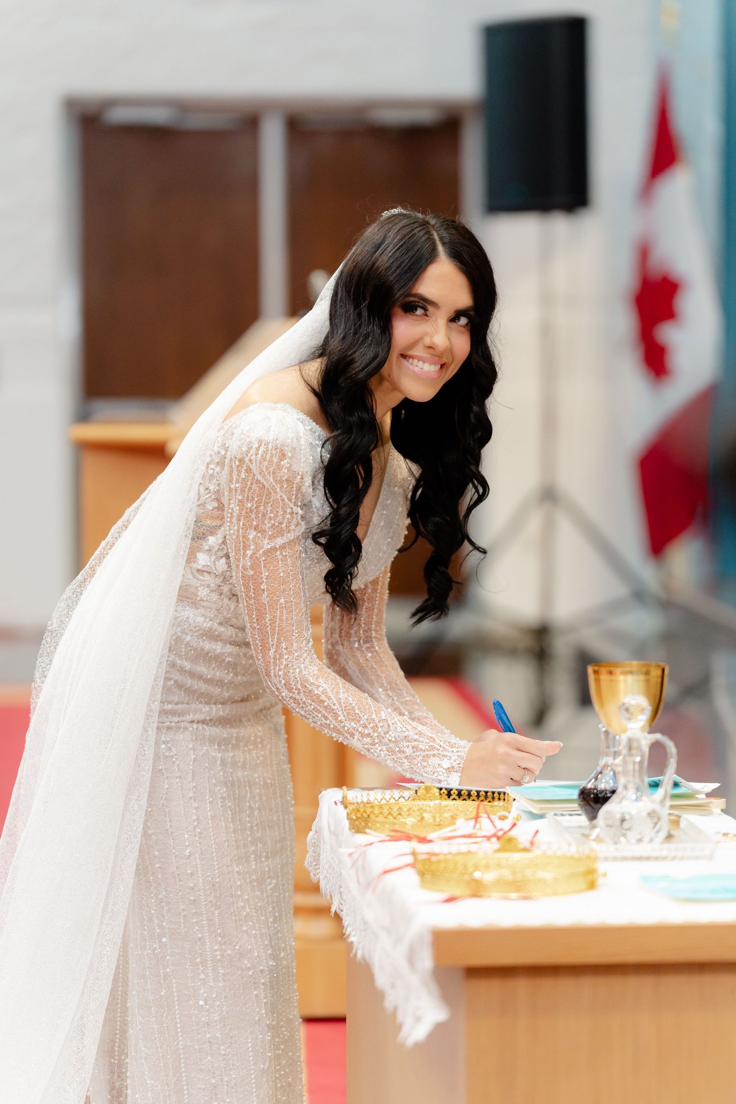 Bride in fitted lace wedding dress smiles while signing documents at ceremonial table.