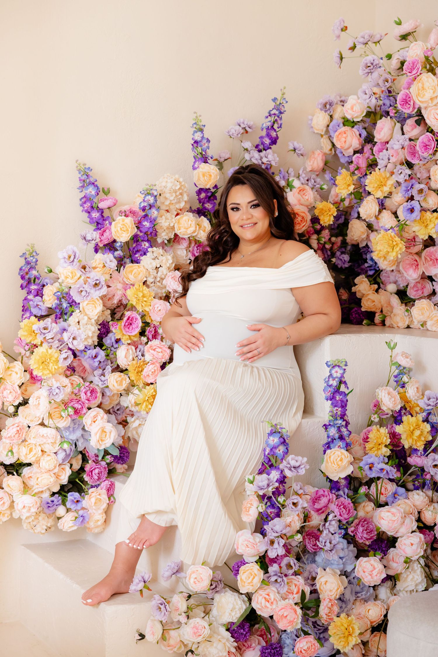 A white maternity gown surrounded by a lush floral arrangement of purple, pink, and yellow blooms against a cream backdrop.