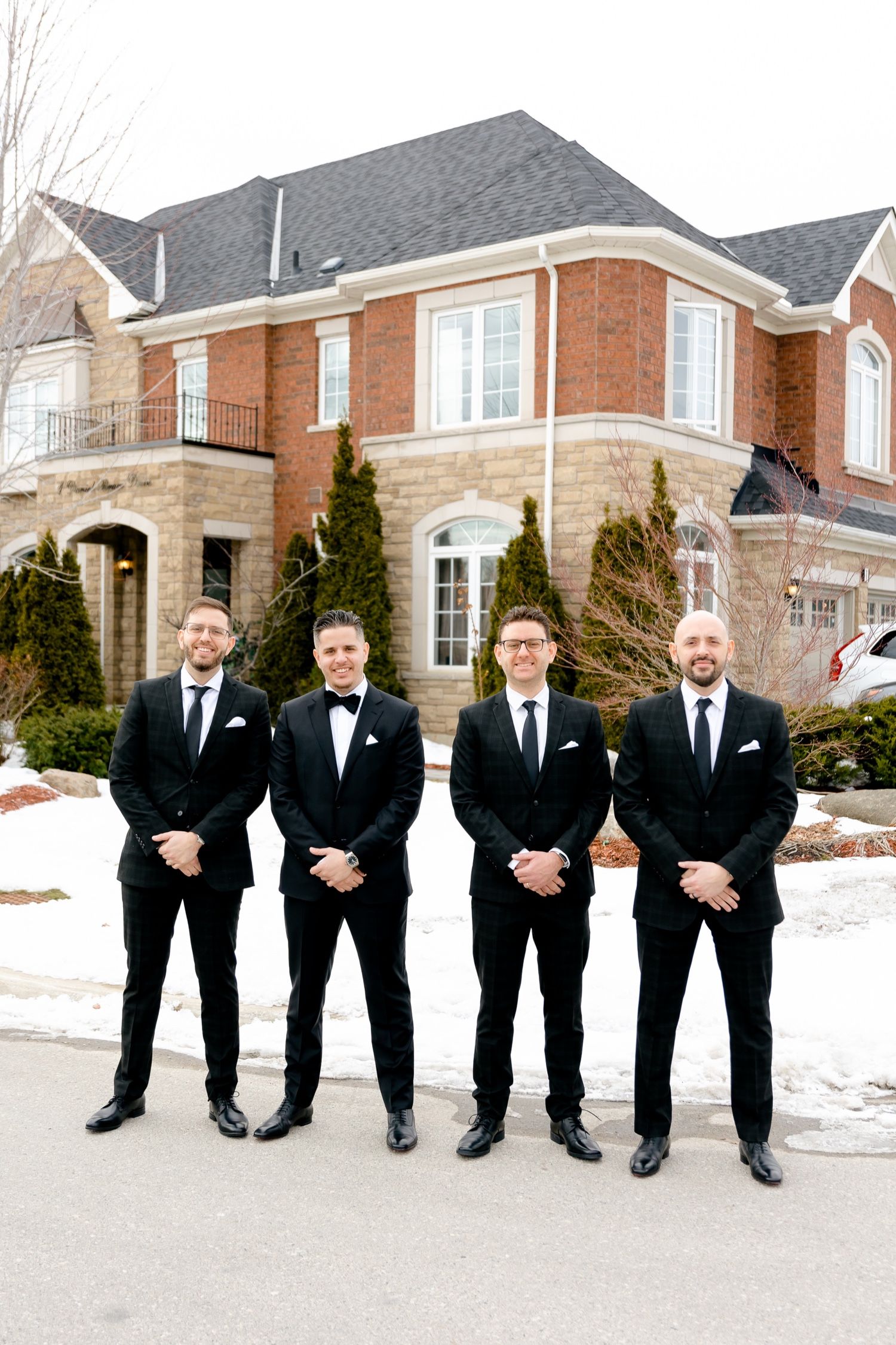 Four groomsmen in black suits pose in front of luxury brick home in winter.