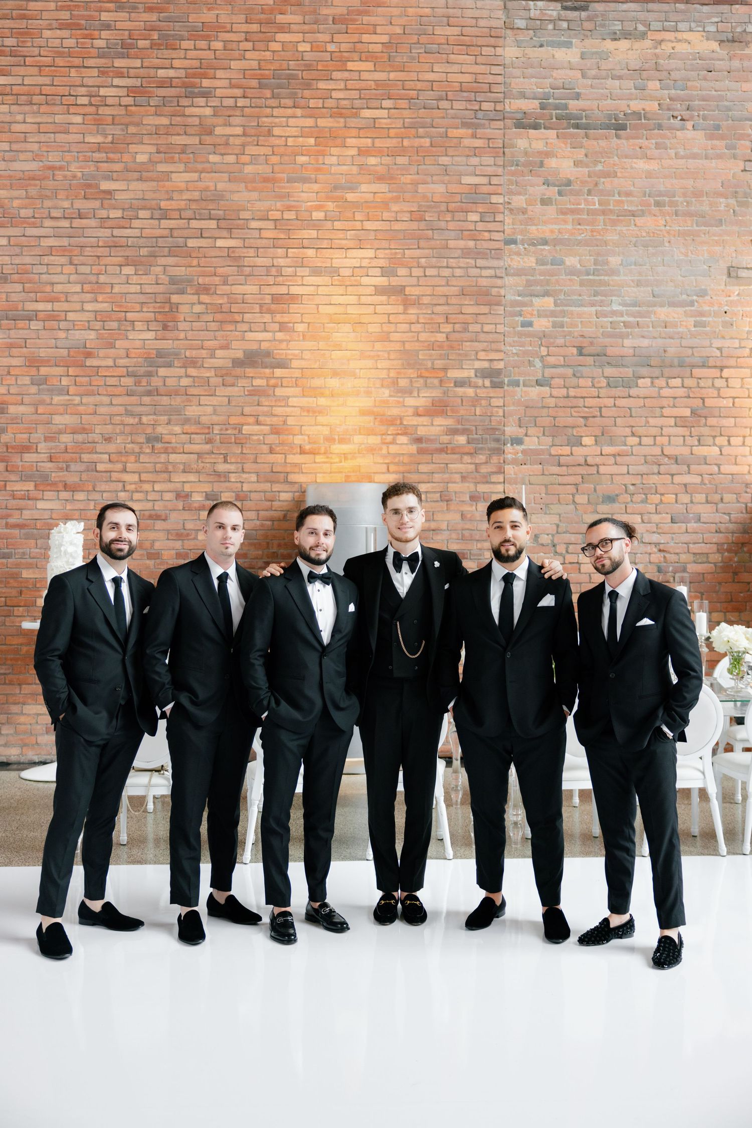 Groomsmen in black tuxedos stand together against exposed brick wall at wedding venue.