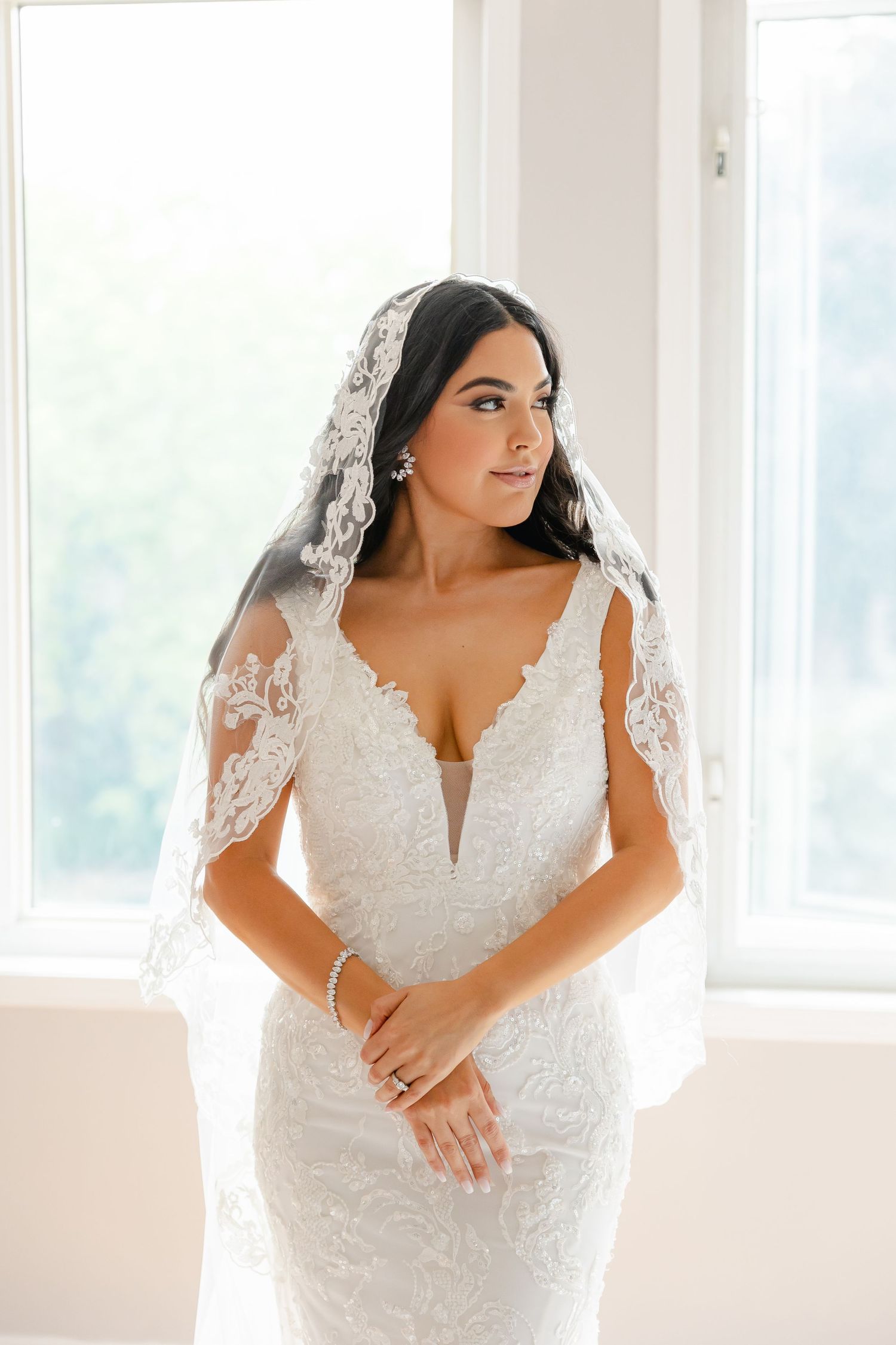 Beautiful bride wearing a fitted lace wedding dress and veil poses by window in soft natural light.