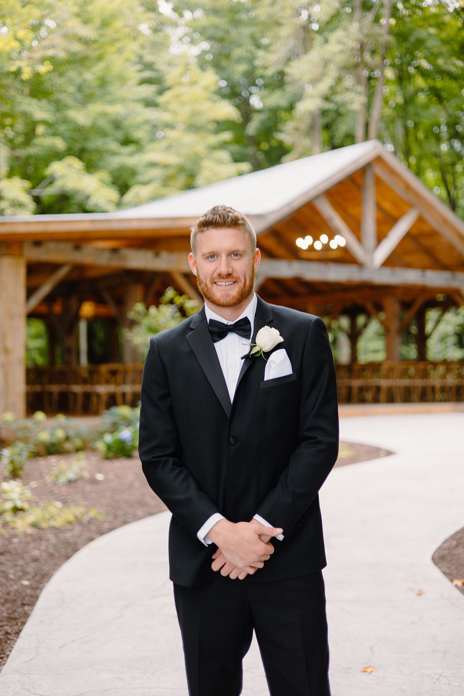 Formal portrait of groom in black tuxedo standing in front of rustic wooden pavilion.