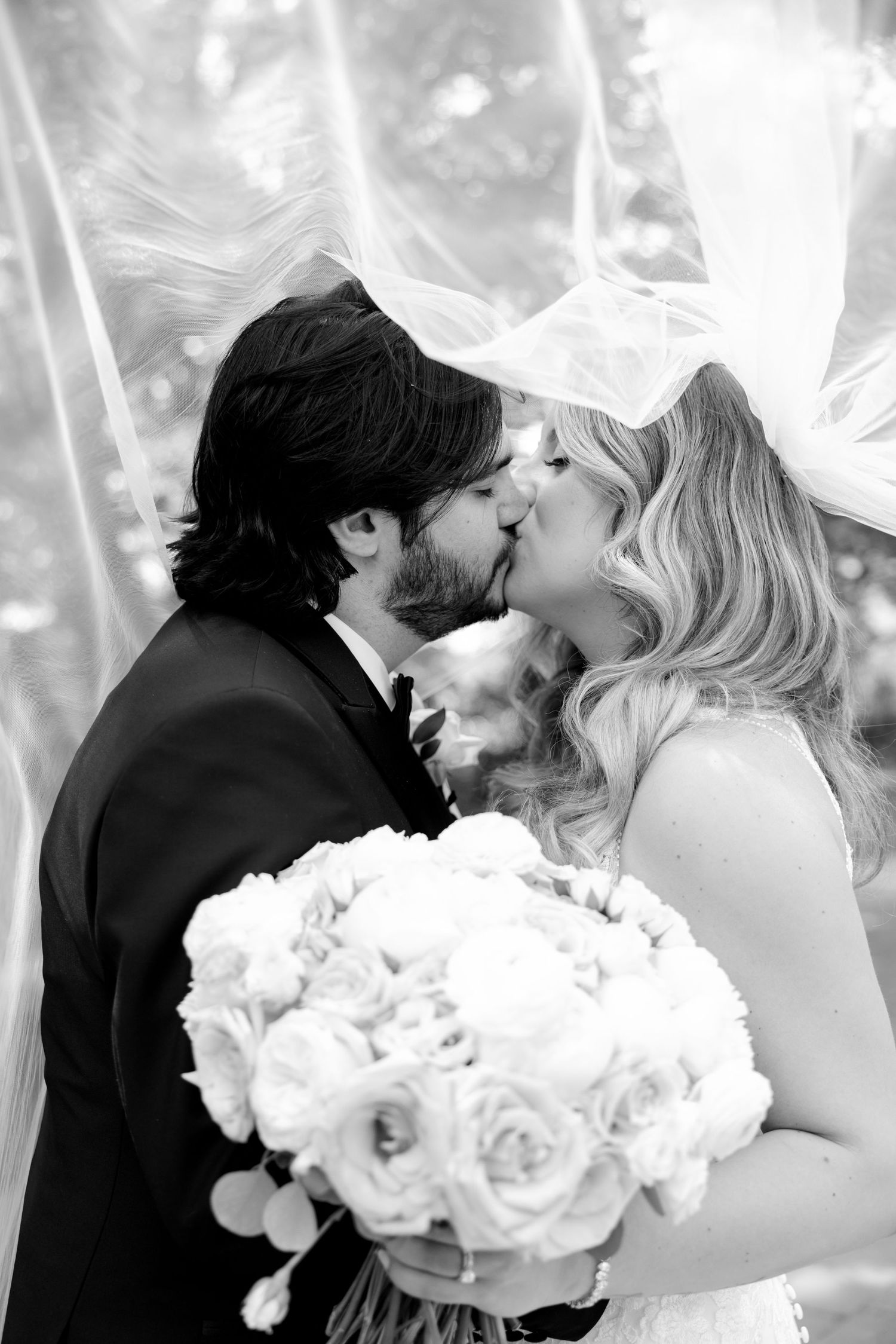 A romantic black and white wedding photo of a couple kissing while holding a bouquet of white roses.