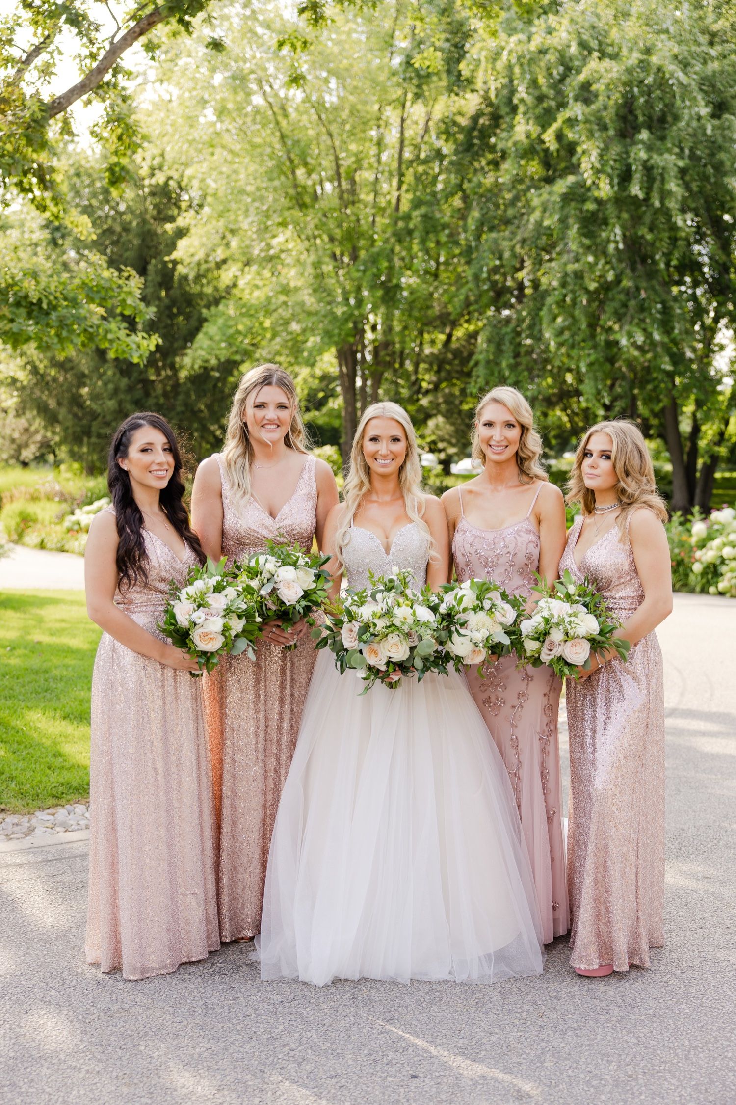 A bridal party in blush pink dresses poses with white floral bouquets on a garden path.