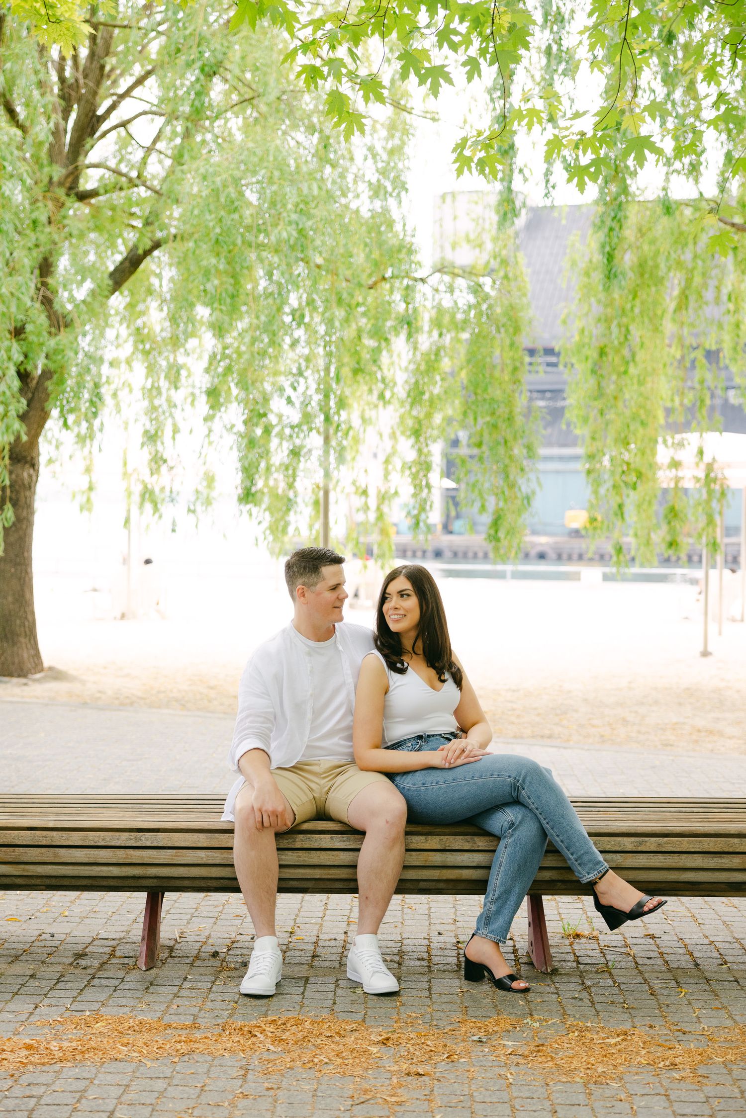 A couple sitting close together on a park bench under lush green willow trees.