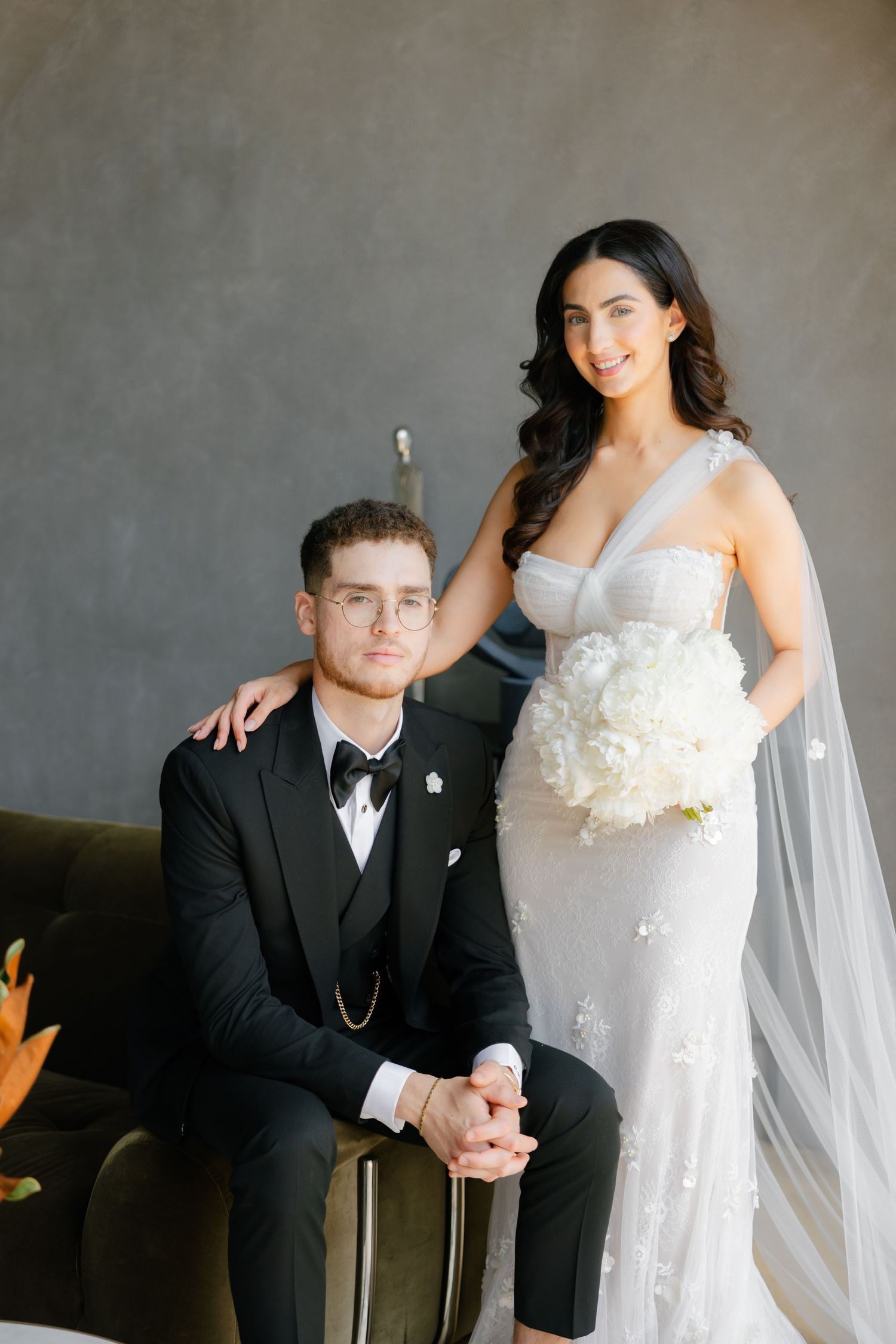 Elegant wedding portrait of couple in formal attire seated on dark couch with floral arrangements.