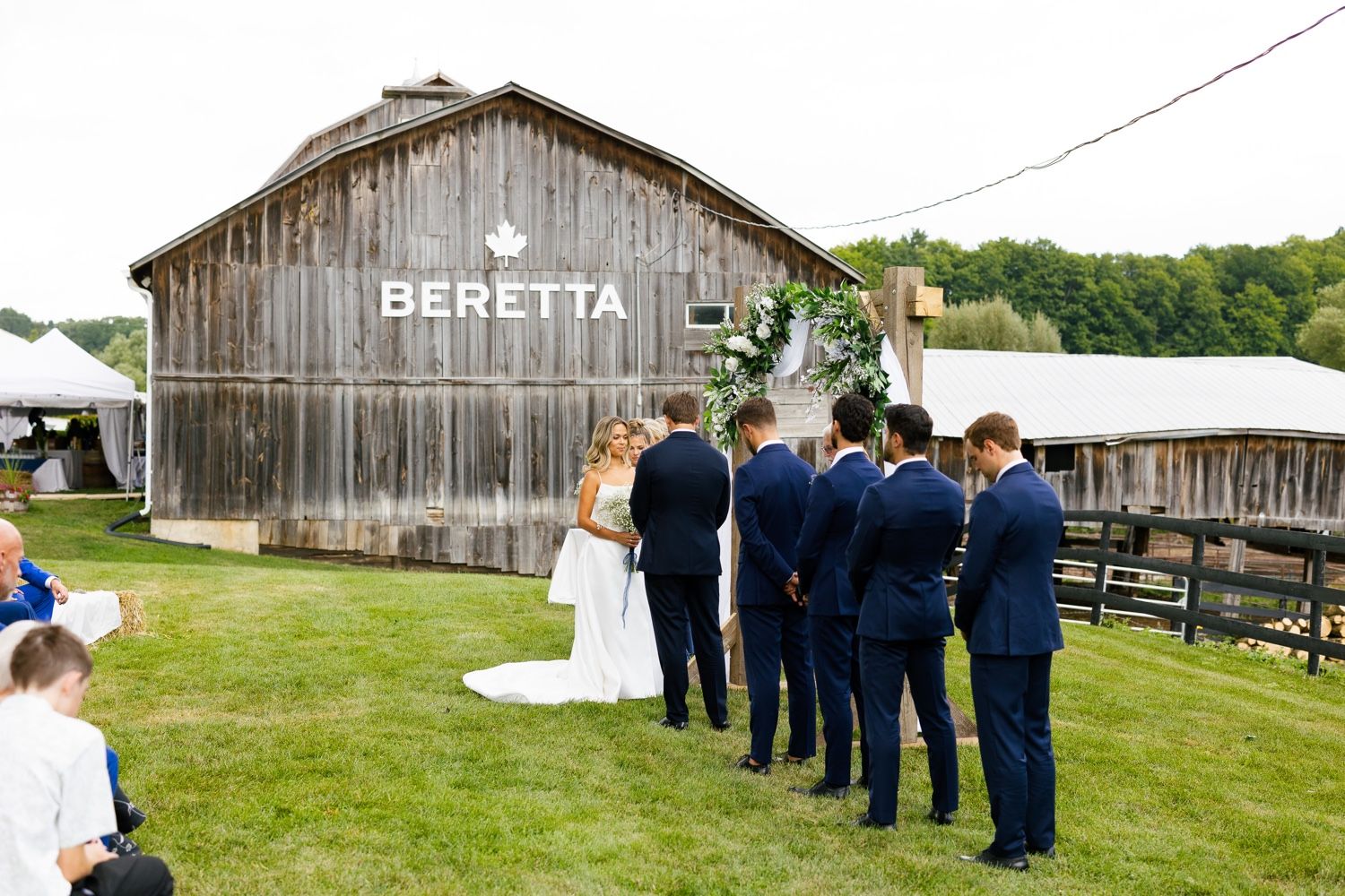 Wedding party poses in front of a rustic wooden barn with 'Beretta' signage.