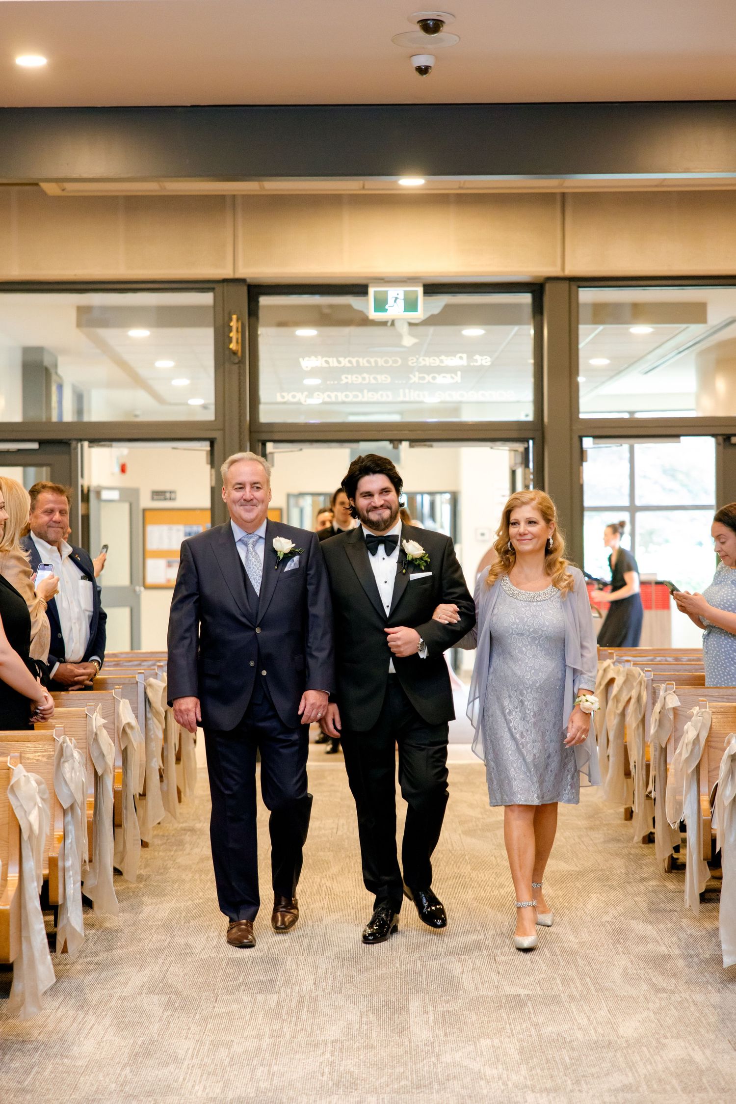 Wedding processional in modern church hallway with wooden pews and glass doors.
