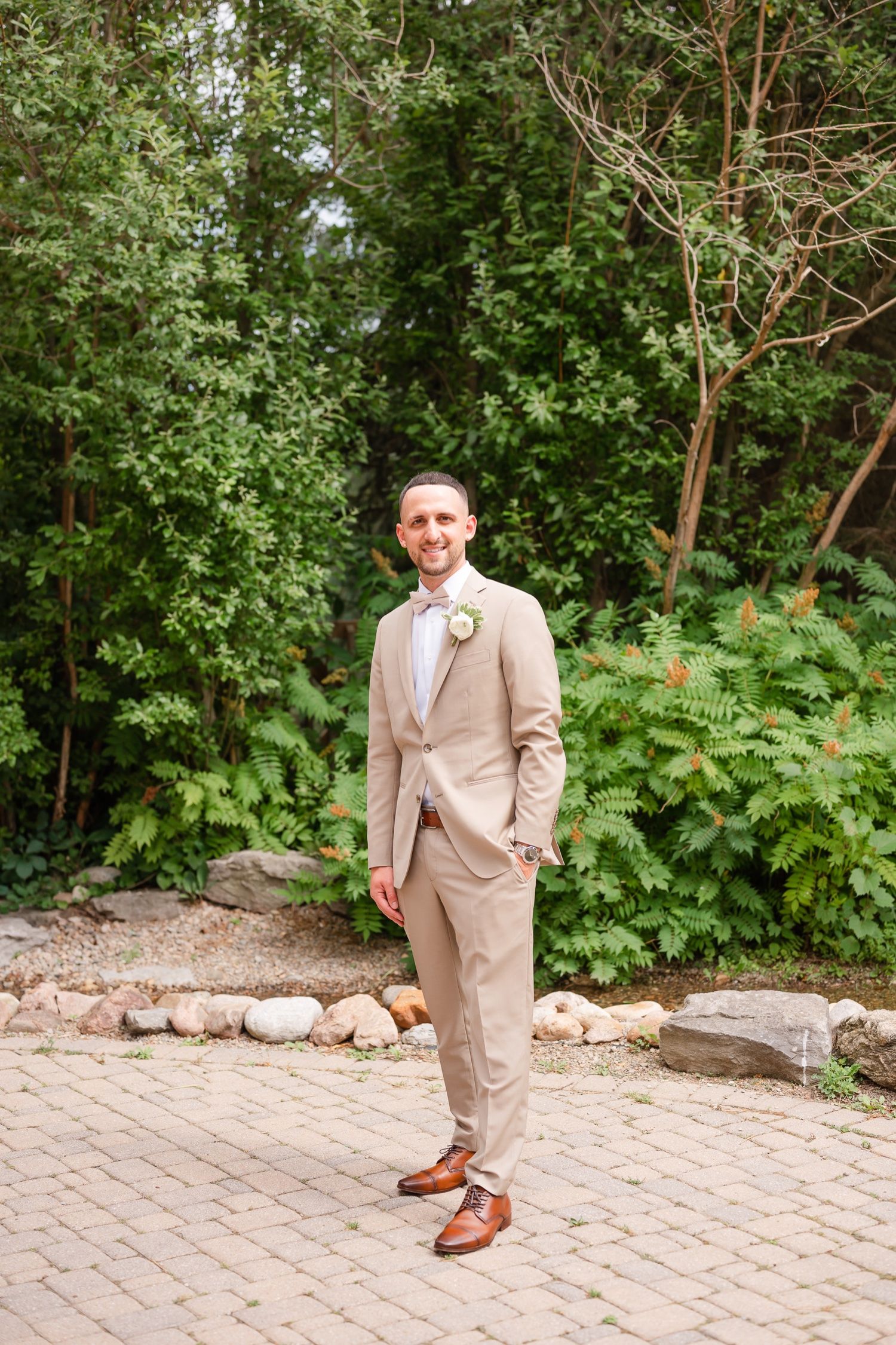 Elegant groom in beige suit and pink bowtie poses on brick paver pathway near garden foliage.