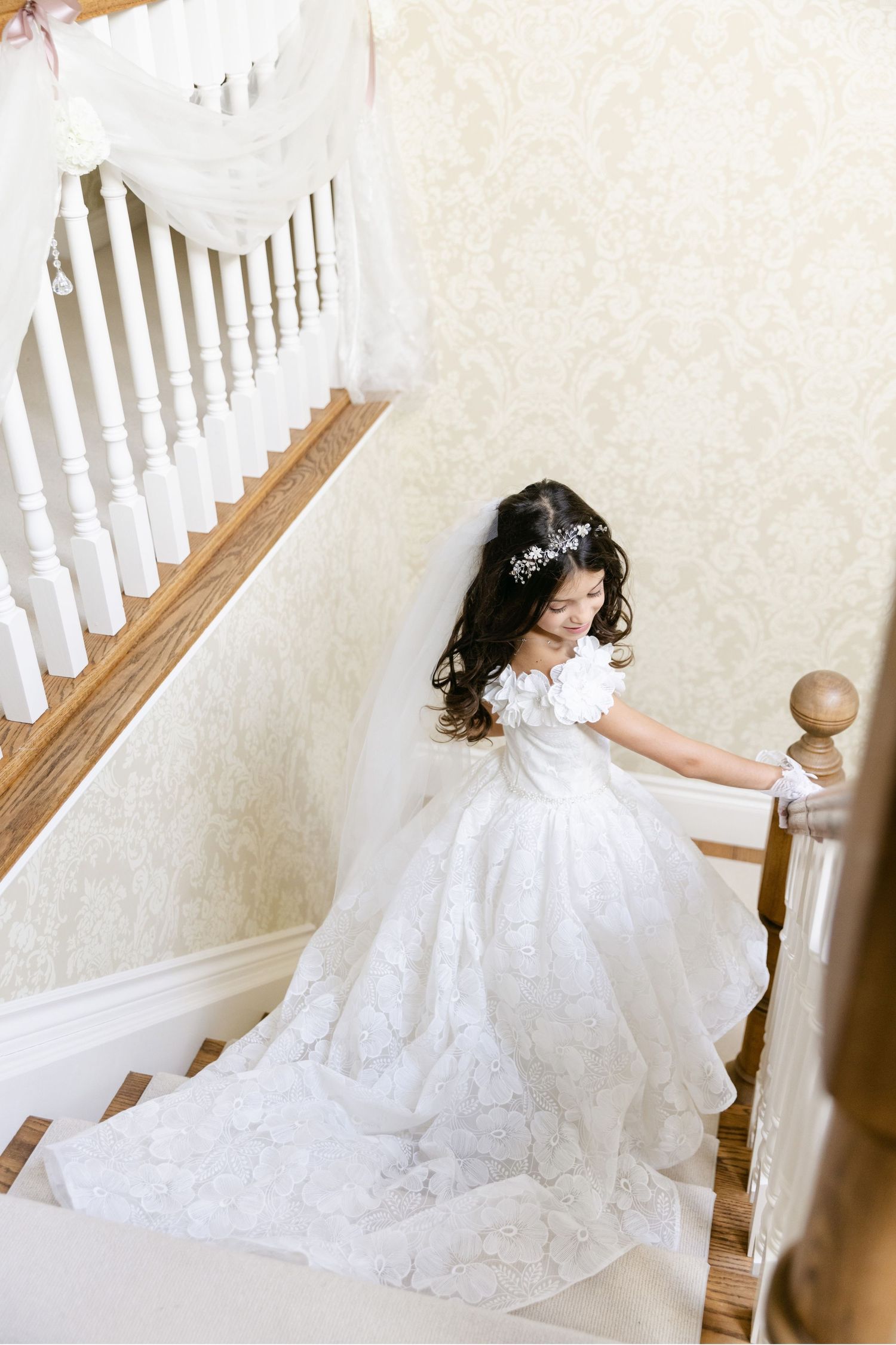 White lace baptism dress with long train flowing down wooden staircase.